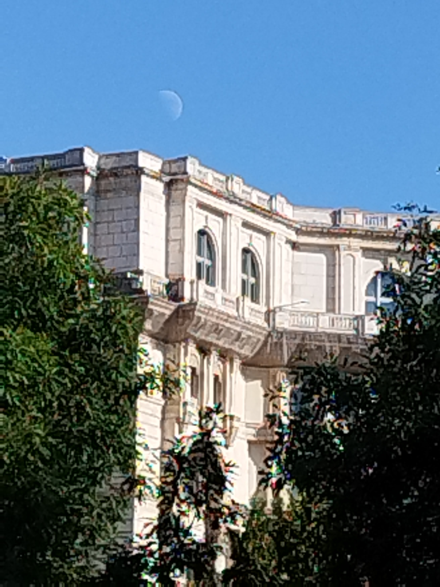 The image shows a crescent moon in a clear blue sky above a building with classical architecture. The building is white with arched windows and decorative moldings. Trees with dark green foliage partially obscure the building. The scene is likely outdoors in a city. The metadata indicates the image was taken in Bucharest, Romania.