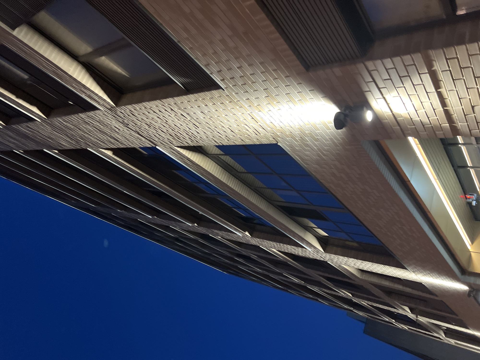 The image depicts the exterior of a brick building at night. The perspective is low-angle, looking up the side of the building against a dark blue sky. The building appears to be several stories high, with numerous windows. Some windows are dark, while others reflect the ambient light or appear to be lit from within. Exterior lights are mounted on the brick facade, casting a bright glow on the wall. The architecture suggests a modern, possibly commercial or residential building. There are no people or visible signs of activity in the frame. The scene is static and focused on the architectural elements of the building. The location is identified as Unknown, Russia.