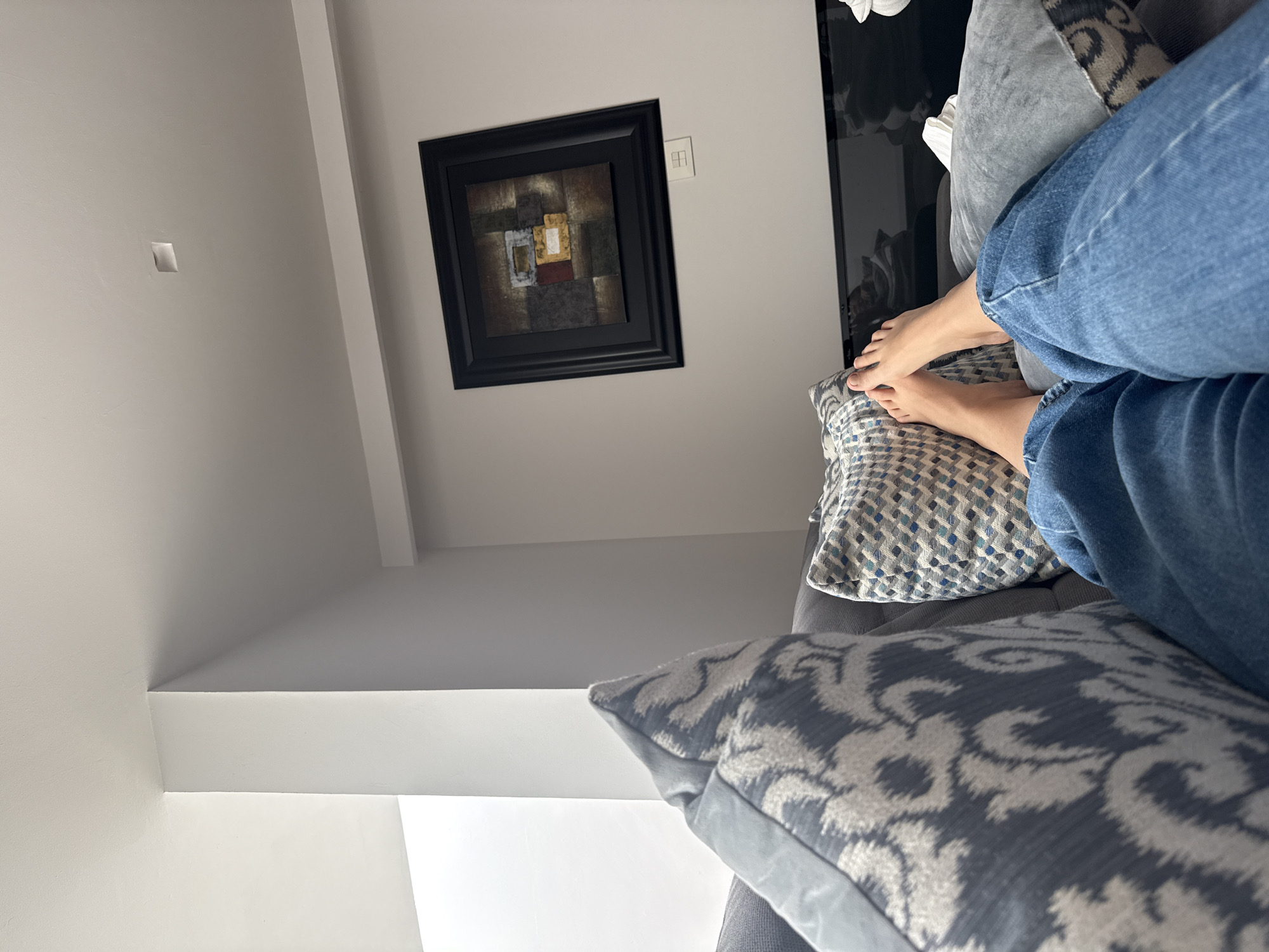 A photograph taken indoors in San Bernardino Tlaxcalancingo, Mexico, shows a person resting on a grey sofa. The person is wearing blue jeans, and their bare feet are visible resting on a blue and white patterned cushion. Other patterned cushions are arranged on the sofa. Behind the sofa is a white wall with a framed, abstract painting and a light switch. The lighting in the room appears to be natural.