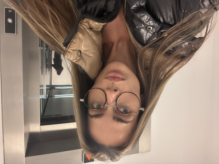 A female subject is depicted from the chest up, taking a selfie in what appears to be an elevator or elevator lobby within the city of Warsaw, Poland. She has long, light brown hair, wears large, round, dark-framed spectacles with a silver bridge, and a glossy black puffer jacket with a visible beige/tan collar. Her lips are slightly parted. To her right, a brushed metal elevator door is visible, featuring a dark square call button and an illuminated red triangular indicator light pointing upwards. The metallic surface reflects a silhouette, consistent with the main subject, appearing to hold a device, confirming the photographic action. The reflection also shows a glimpse of an interior space beyond the subject, possibly the elevator cabin or an adjacent hallway.
