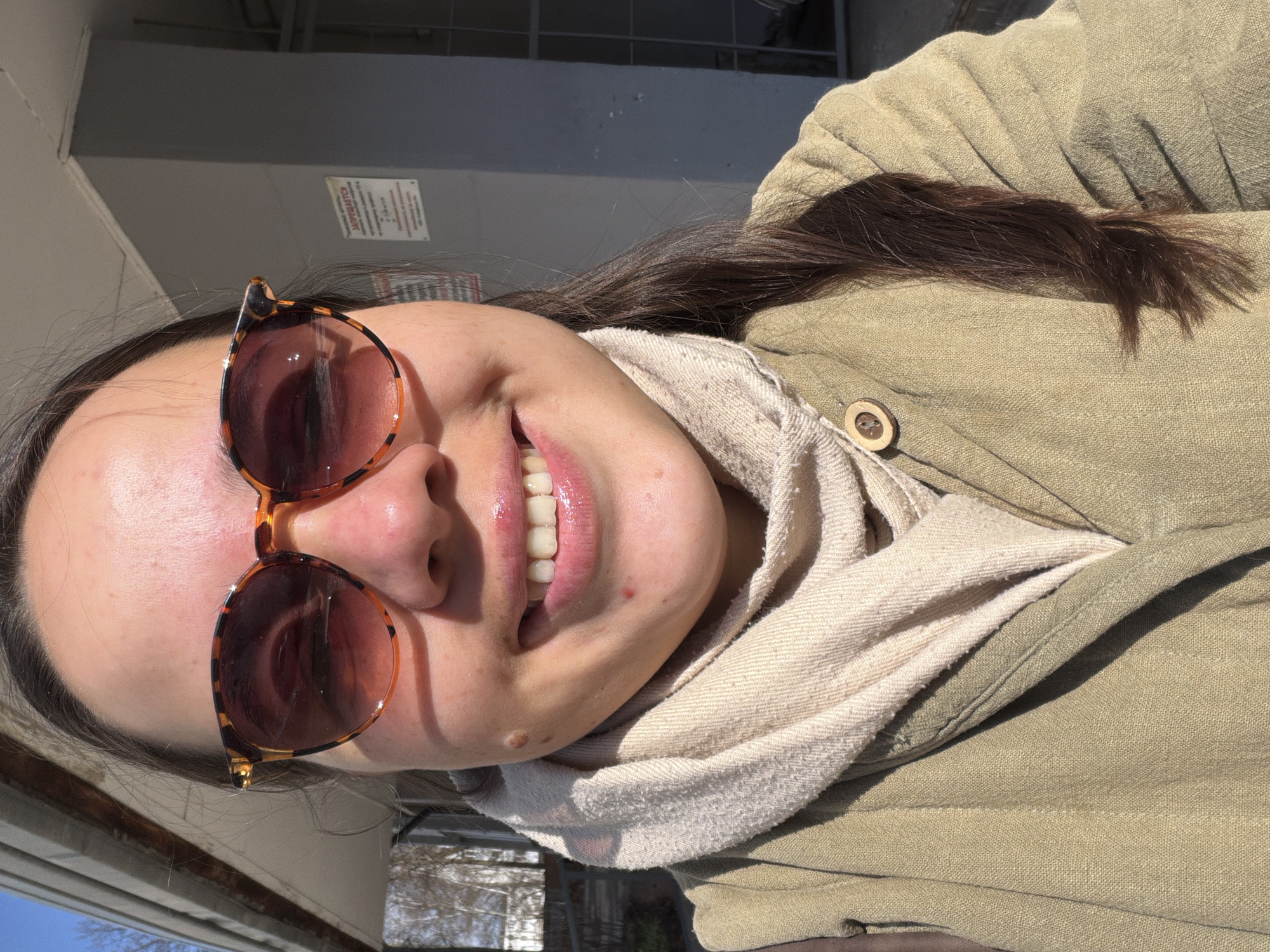 A young woman is captured in a brightly lit, close-up outdoor portrait, possibly a selfie. She is smiling broadly, revealing her upper teeth. Her dark brown hair is long and falls over her shoulders. She wears tortoiseshell-patterned sunglasses with dark brown lenses, partially covering her eyes. Around her neck is a light, possibly cream or beige, textured scarf. Over it, she wears a khaki or olive-green jacket or coat made of a textured fabric, featuring a visible wooden button on the front.
The background is somewhat blurred but indicates an outdoor, urban or semi-urban setting under clear, sunny skies, suggesting midday or early afternoon. A gray building or wall is visible behind her, with a white rectangular sign affixed to it. The most prominent visible text on this sign is the Russian word "ЗАПРЕЩАЕТСЯ" (ZAPRESHCHAYETSYA), meaning "FORBIDDEN" or "PROHIBITED." Hints of other structures or distant elements can be seen in the lower left background. The overall mood is cheerful and relaxed. This image is set in Chaikovsky, Russia.