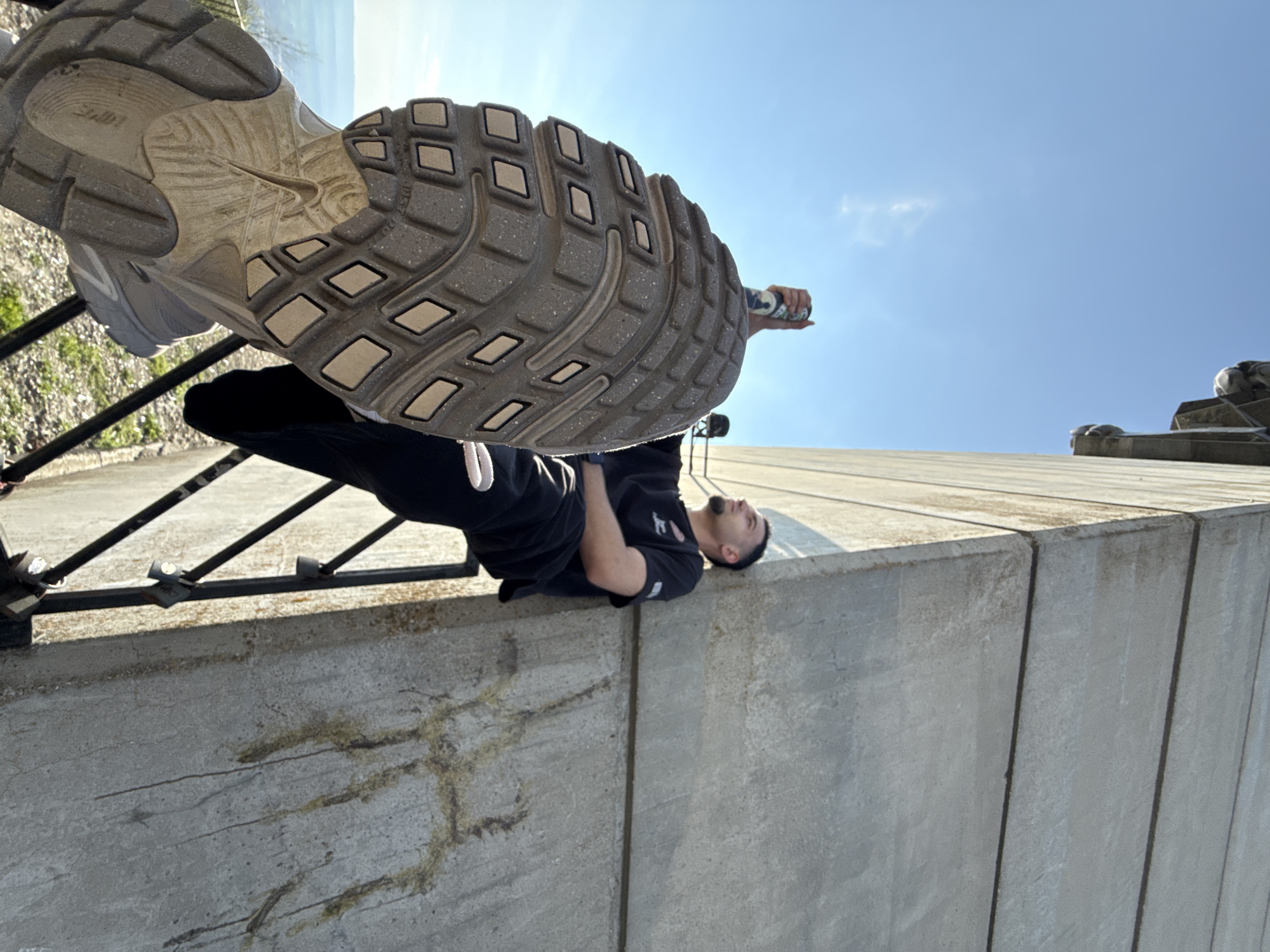 A low-angle shot shows a man lying on a concrete ledge, with the sole of a sneaker obscuring part of the view. The ledge has a metal railing behind the man. He is wearing a black t-shirt and black pants. He looks upwards with his head tilted back. In his right hand, he appears to be holding a can of spray paint. The sky is clear and blue. Part of the Monument to 1300 Years of Bulgaria is visible to the right, which places the location in Shumen, Bulgaria.