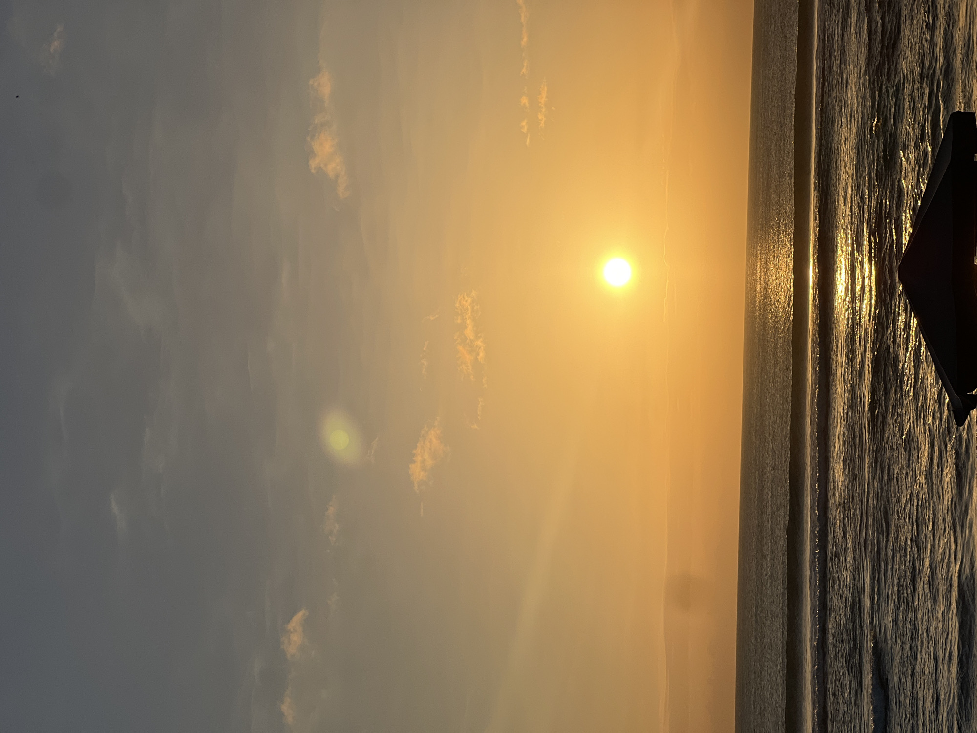 The image depicts a serene sunset over the ocean in Kuta, Indonesia. The sun, a bright orb, is positioned in the center of the frame, casting a warm golden glow across the sky and reflecting shimmering light onto the water. Wispy clouds are scattered across the sky, some tinged with orange and pink from the setting sun. Gentle waves roll towards the shore, creating a tranquil atmosphere. In the foreground, a dark silhouette of a structure, possibly a beach umbrella or a small pavilion, is visible at the bottom edge of the image, providing a point of contrast against the luminous sky and sea. The overall scene conveys a sense of peace and natural beauty.
