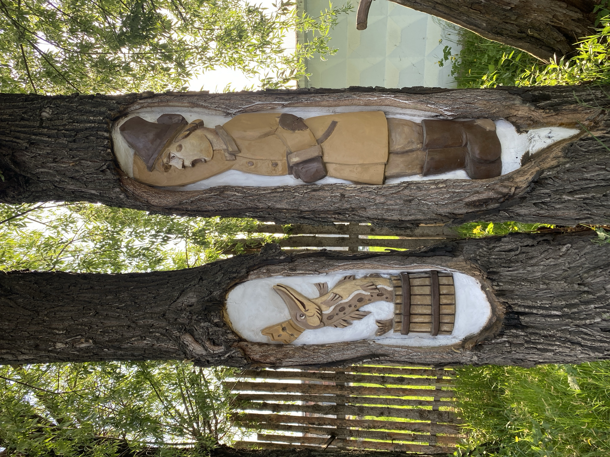 Two tree trunks have carvings in them. The top carving depicts a man lying down, wearing a hat, a jacket, and boots. The bottom carving shows a bird-like figure beside a fish, with both next to a barrel. The carvings are set in oval recesses in the tree trunks and are painted with white backgrounds. Foliage and a fence are visible in the background. This scene is located in Chaikovsky, Russia.