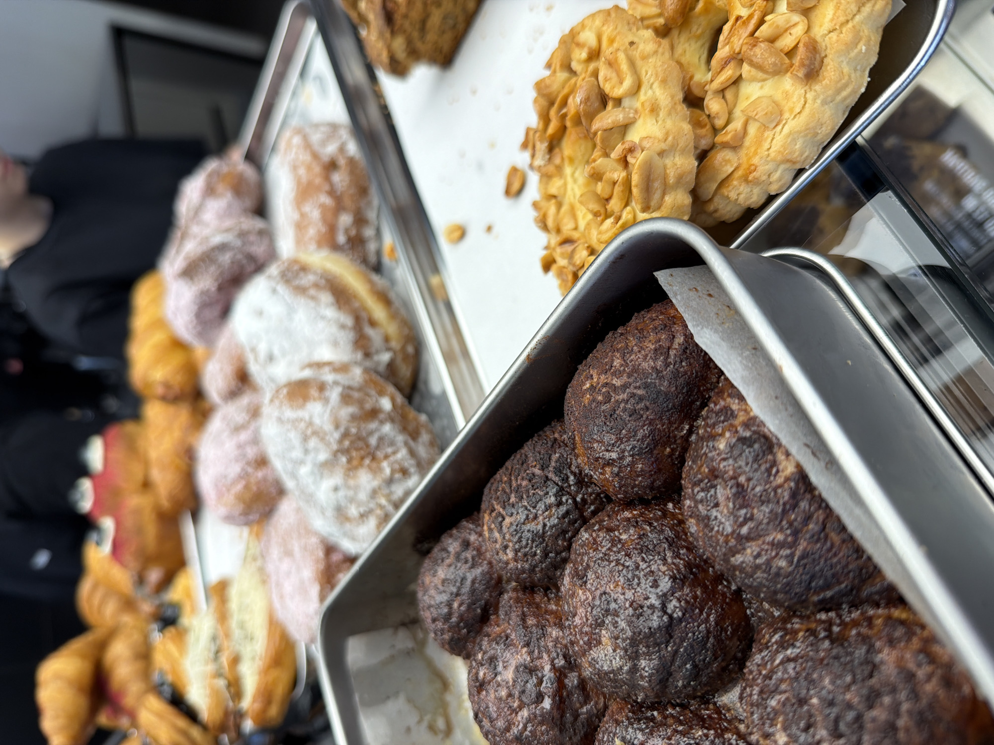A close-up shot displays a selection of pastries in metal trays. The top tray contains cookies adorned with peanuts, and a sliced cake. The lower tray features small, dark, round pastries. Additional pastries are visible in the background, including croissants and powder-covered doughnuts. The location and city are not identifiable from the given image.
