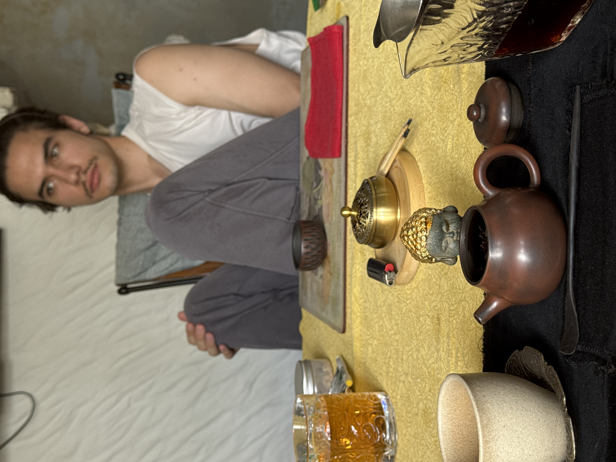 A man sits cross-legged behind a table draped in a yellow patterned cloth. The table displays tea-related objects, including a teapot, a teacup, a glass pitcher, an incense burner, a lighter, and a small Buddha statue. The location cannot be determined based on the image.