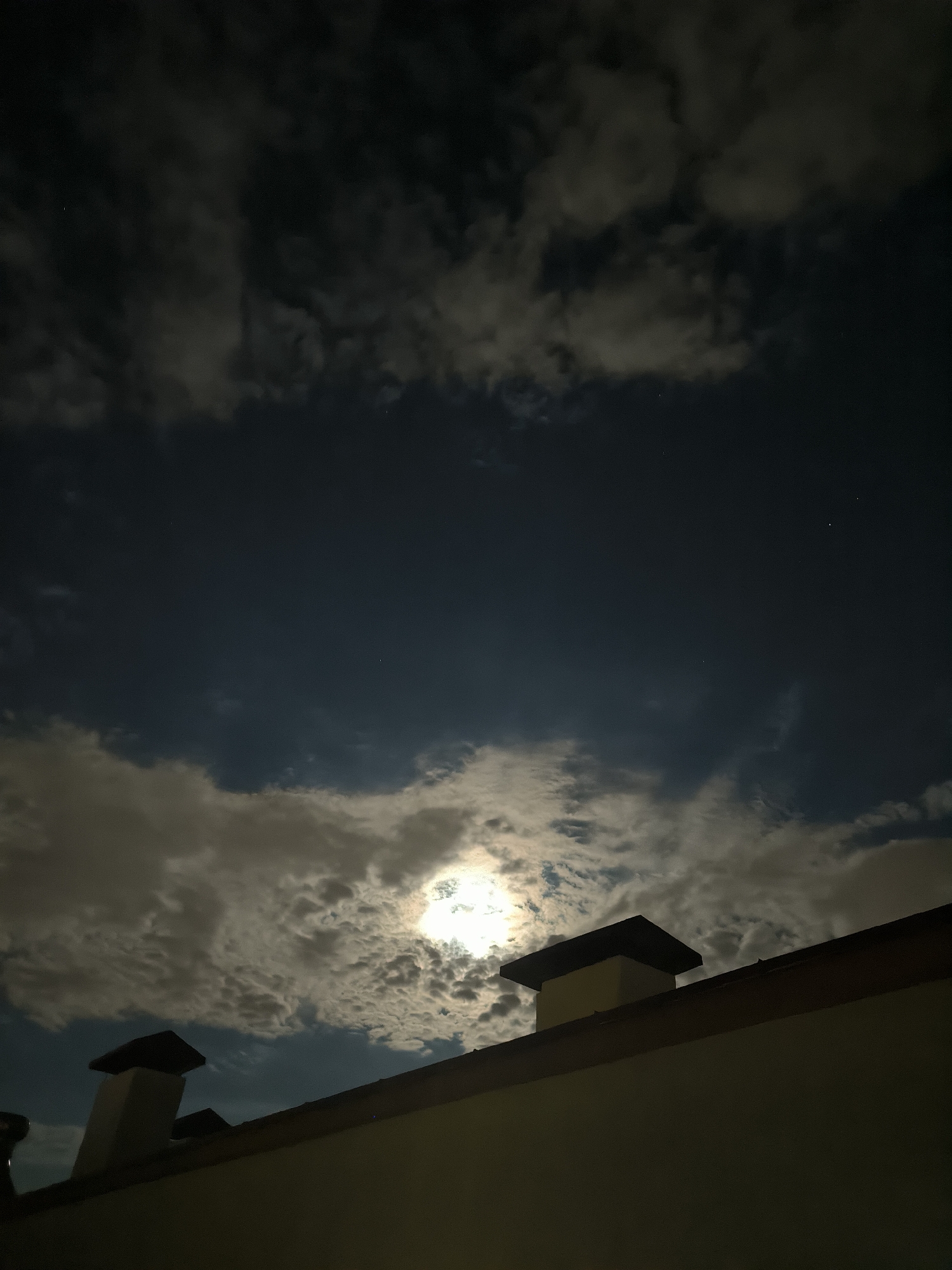 The image shows a night sky with clouds illuminated by the moon. Two chimneys and a portion of a roof are visible in the foreground, silhouetted against the sky. The moon is partially obscured by clouds, creating a dramatic effect. The scene appears to be from a residential area, possibly a rooftop view in Chișinău, Moldova. There are no people or specific actions depicted.