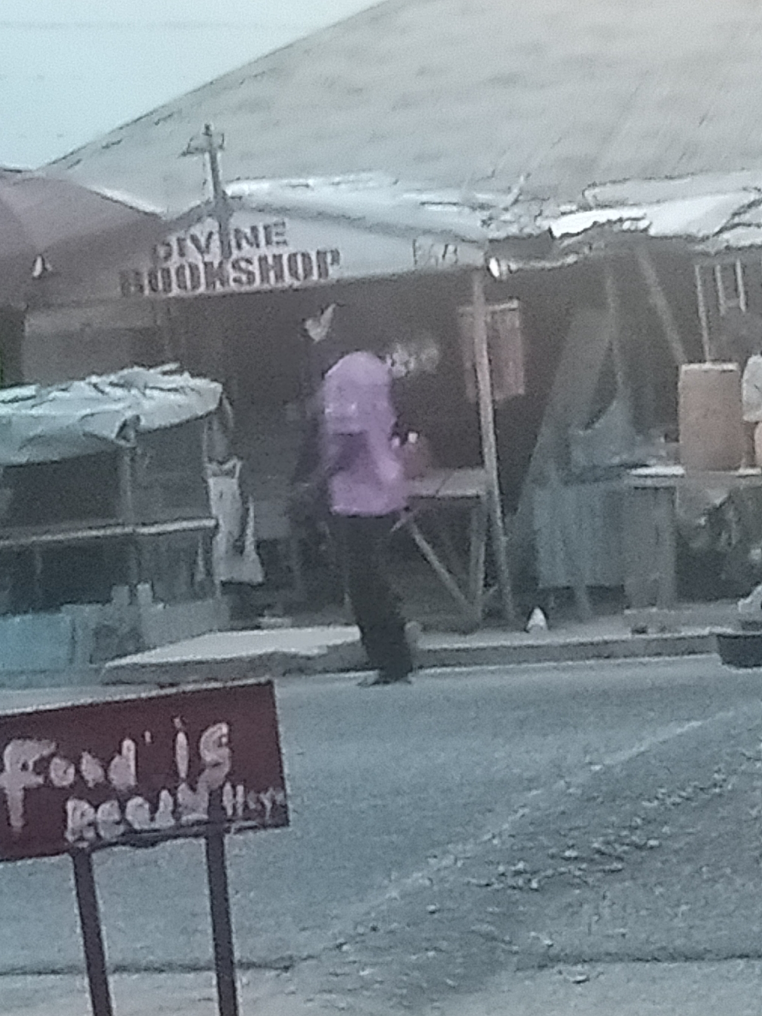 The image depicts a street scene in Jalingo, Nigeria. A building with a sign reading "DIVINE BOOKSHOP" is visible. A person wearing a purple shirt is standing in front of the building. A red sign in the foreground says "Food is Ready". The environment appears to be an outdoor market or commercial area with stalls and structures.