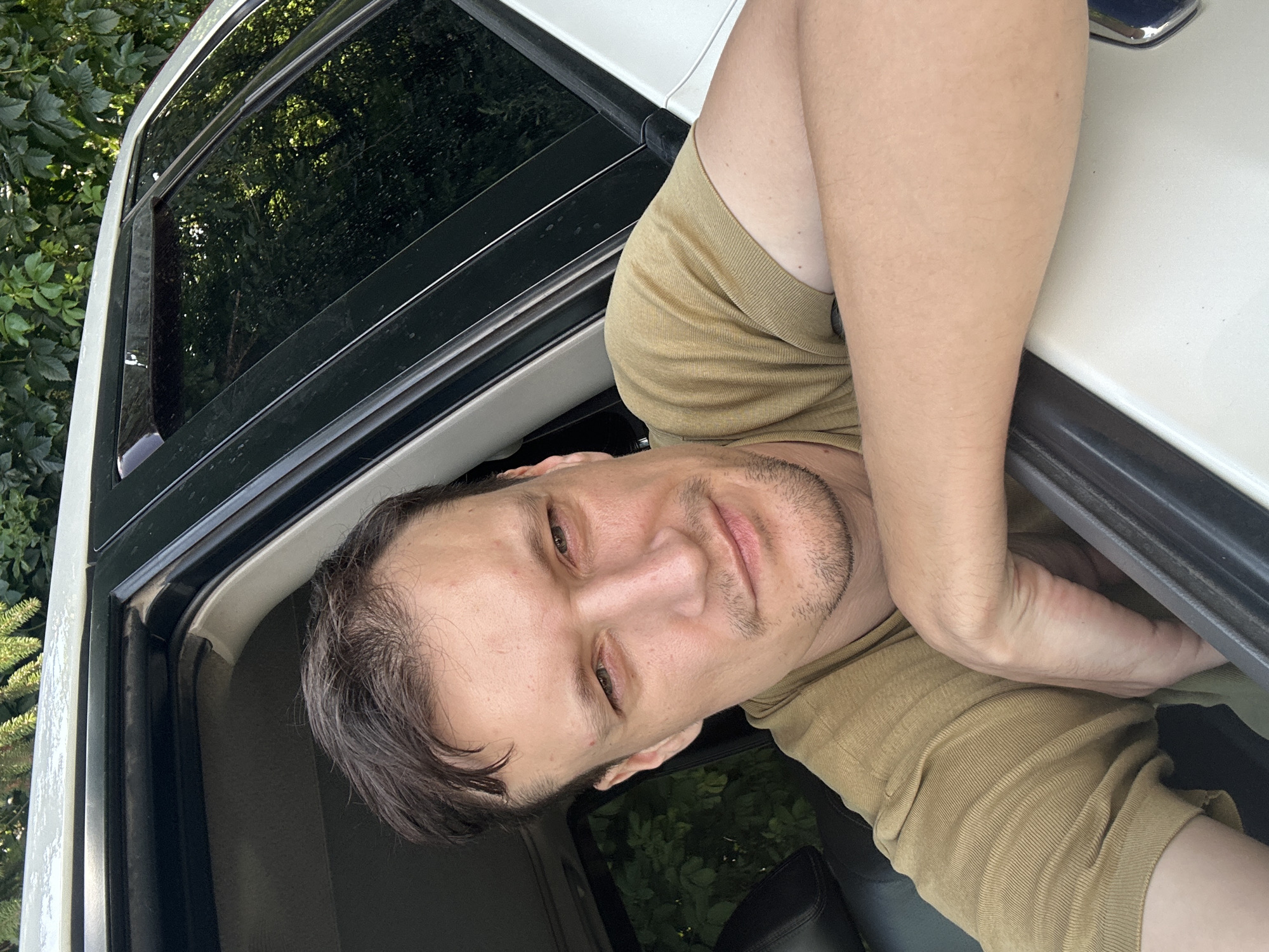 In Shatava, Ukraine, a man with short, dark brown hair and light skin is captured in a selfie taken from inside a vehicle. He is leaning on the open car door, with his right arm resting on the door frame and his hand draped over the top. The man is wearing a plain, olive-green t-shirt with a ribbed texture. His head is tilted slightly, and he is looking directly at the camera with a neutral expression. His facial features include a prominent nose, thin lips, and a light stubble on his chin and upper lip. A small, reddish mark is visible on his forehead, and his skin appears slightly flushed. The interior of the car shows a dark leather seat behind him and the car's window frame with tinted glass. Outside the car, lush green foliage is visible, suggesting the vehicle is parked outdoors in a natural setting. The car is white, and a portion of its roof is visible at the top of the frame, with some faint reflections on its surface.