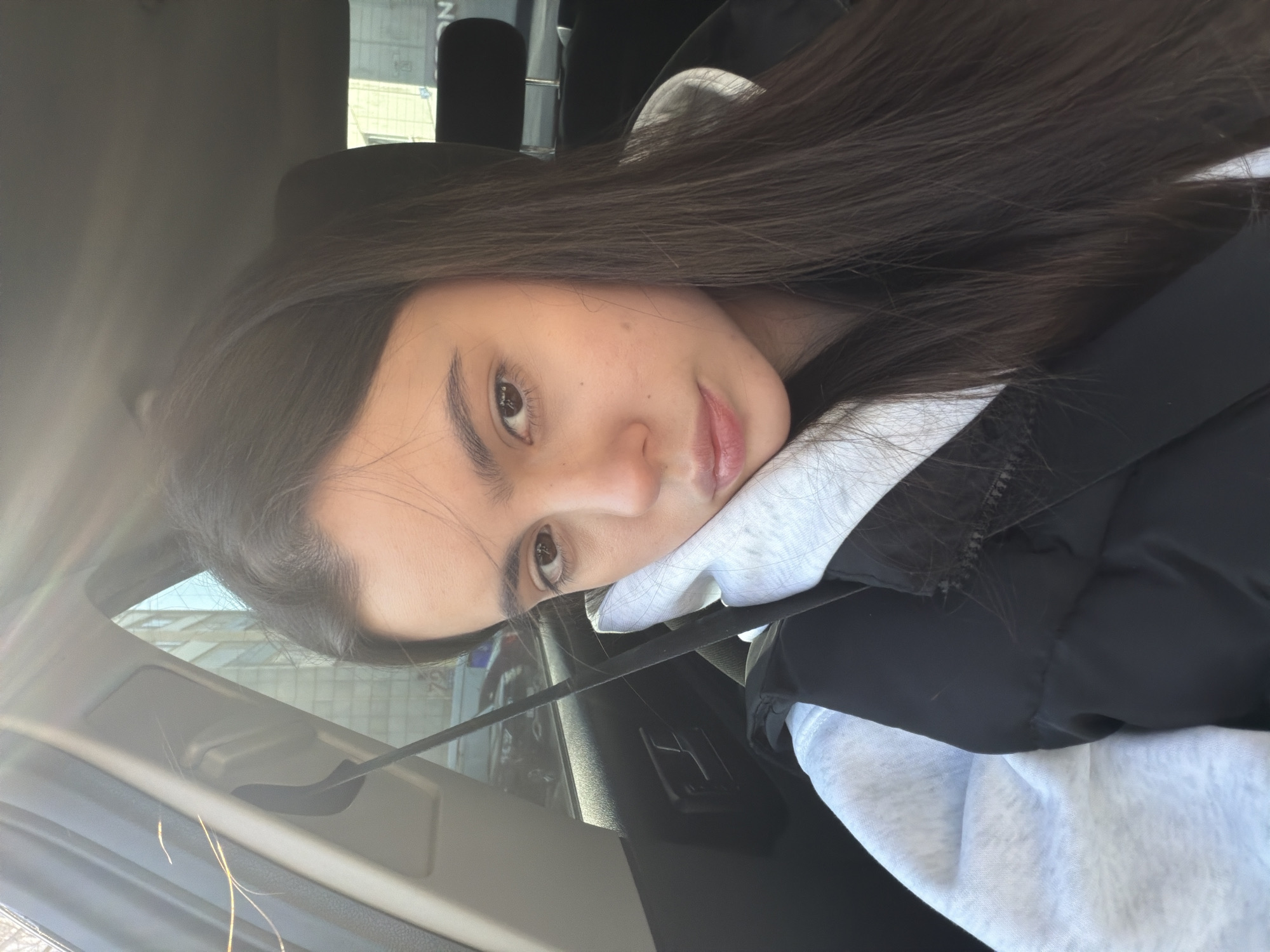A young woman with long, dark brown hair is sitting in a car. She is wearing a black vest over a light gray hooded sweatshirt. Her face is slightly angled towards the camera, and she appears to be looking directly at the lens. The interior of the car is visible, including the dashboard and the seatbelt. The exterior view through the window suggests an urban setting, possibly in Chaikovsky, Russia, though specific landmarks are not discernible. The light suggests it is daytime.