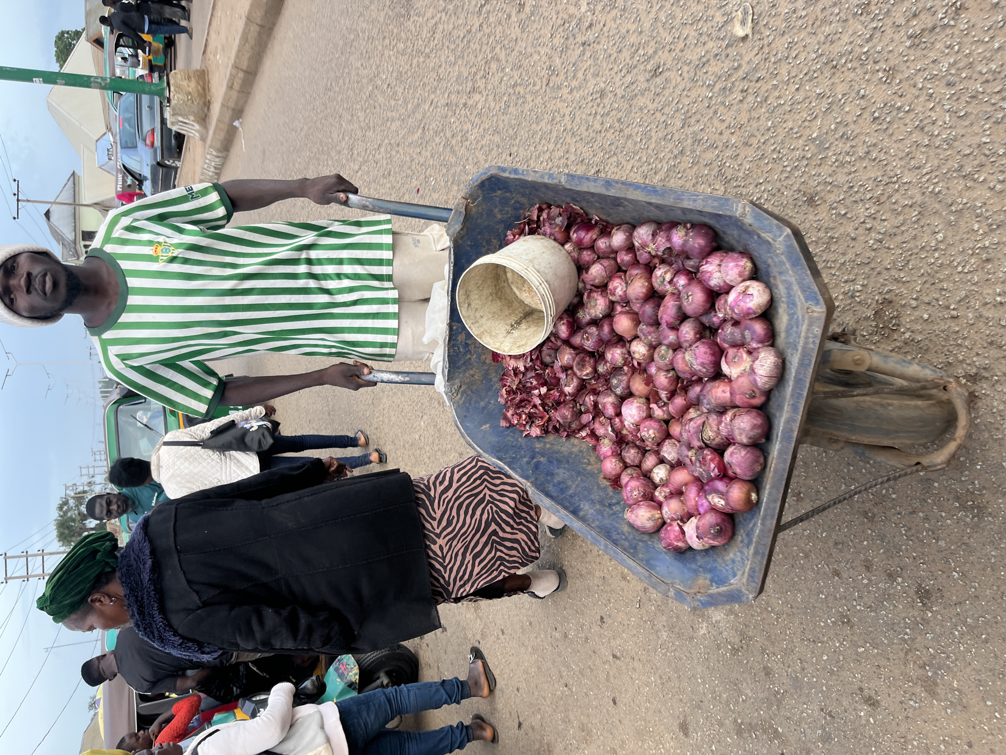 A street vendor pushes a wheelbarrow full of red onions in Bukuru, Nigeria. The vendor, a Black male wearing a white beanie and a green and white striped t-shirt, grips the handles of the wheelbarrow. The wheelbarrow is blue and contains a large quantity of red onions, along with a white plastic bucket. In the background, other people are visible, including a woman in a green headwrap and black coat, and a person in a white jacket. Several vehicles, including a green auto-rickshaw and a grey car, are also present on the street. The ground is paved with asphalt. The sky is overcast.