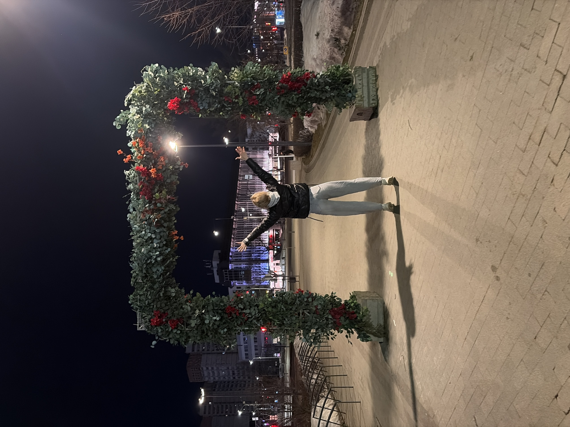 A nighttime scene in Perm, Russia, depicts a single individual standing with their back to the camera, arms raised and outstretched, centered beneath a decorative archway. The person has light-colored hair, possibly covered by a hood, and wears a dark, presumably black, puffer-style jacket, light gray or light-wash pants, and light-colored athletic shoes. Their posture casts a long shadow extending forward on the paved ground.
The archway, the primary focal point besides the person, is densely covered with dark green foliage and clusters of bright red artificial flowers or berries. It is supported by two vertical pillars, each emerging from a rectangular concrete-like base resting on the ground.
The environment is an urban setting, illuminated by artificial light sources. The ground is paved with rectangular blocks, and patches of snow or ice are visible on the periphery. In the background, multiple multi-story buildings are visible, some brightly lit from within and by exterior lighting. Streetlights are prominent throughout the scene, casting distinct shadows. Bare tree branches are visible on the right, and a row of bicycle racks is present on the left. A pole displaying the Cyrillic text "СТО" is visible in the left midground. Distant traffic lights and other illuminated signs suggest an active urban street beyond the immediate foreground.