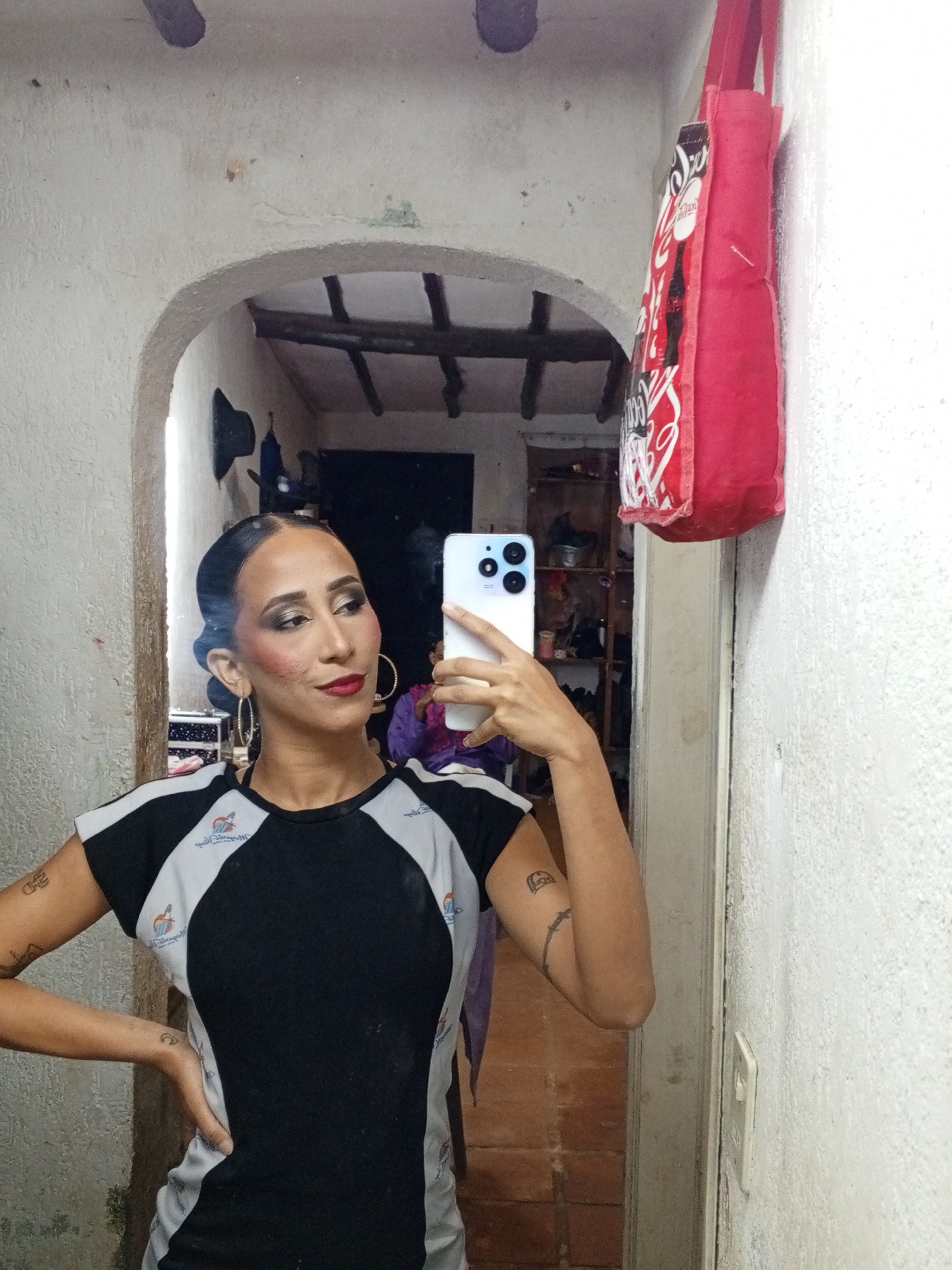 A woman with dark hair pulled back, hoop earrings, and visible makeup (dark eye shadow, red lipstick, and rouge) is taking a selfie in a mirror using a white cell phone. She is wearing a black and grey t-shirt. She is standing in a hallway with an arched doorway visible behind her. A red Coca-Cola tote bag is hanging on the wall to the right of the mirror. The hallway appears to lead to another room with shelves and various objects. The location is Porlamar, Venezuela, based on visible details.