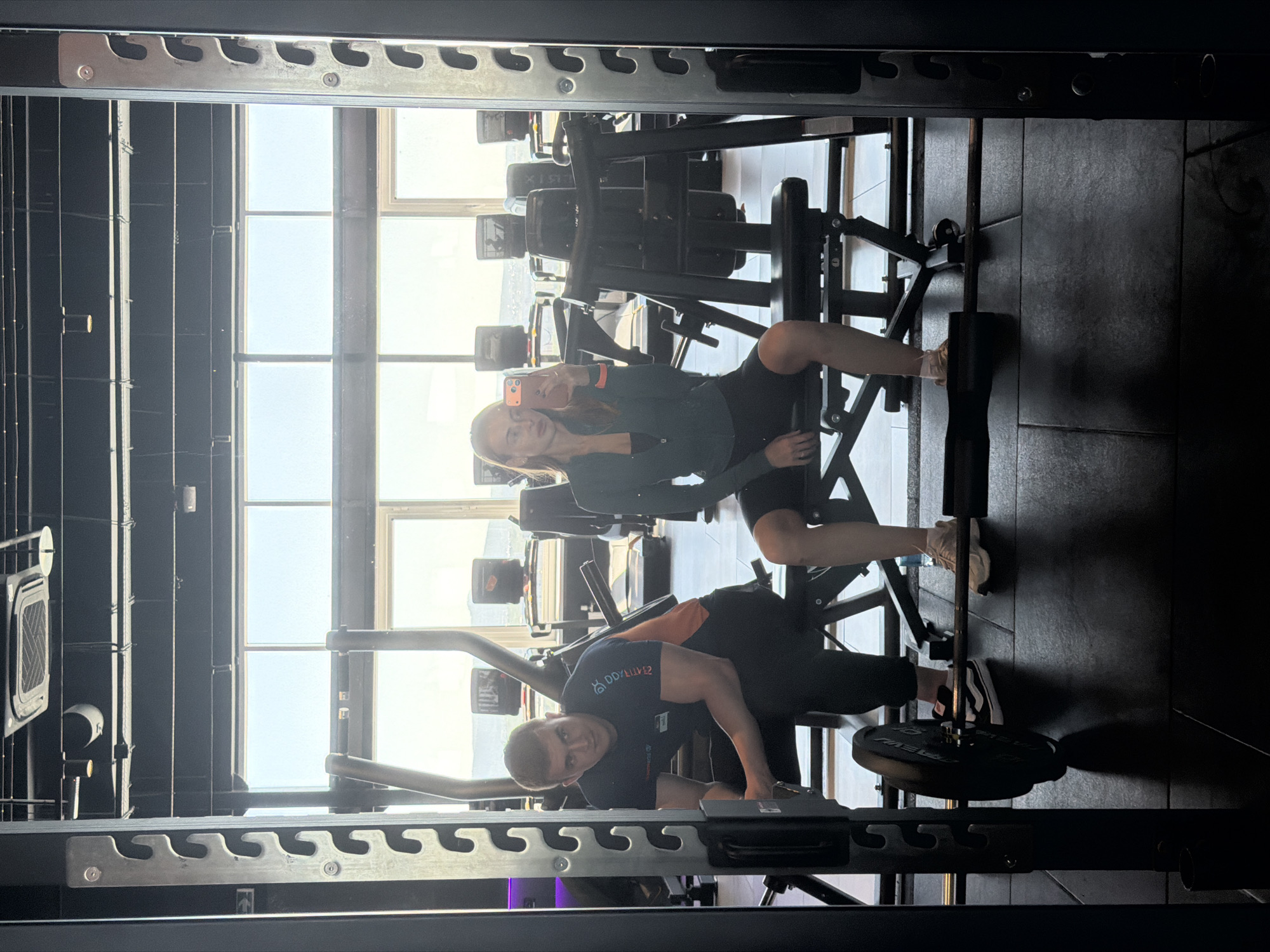 This image captures two individuals, a woman and a man, inside a modern gym, taken as a mirror selfie. The woman is seated on a weight machine on the right side of the frame, holding a smartphone to take the picture. She has long, light brown hair and is dressed in a dark top and dark shorts.
On the left side of the frame, a man is positioned on the floor, leaning over another piece of gym equipment, appearing to adjust weights or prepare for an exercise. He wears a dark t-shirt featuring a blue and red logo and the text "DDULUV2" with "PRO FORM" partially visible underneath. He is looking towards the camera. A black weight plate with "PRECOR" inscribed on it is visible near his feet.
The gym features various dark-colored weight machines and structural metal racks. Bright natural daylight floods the space through large windows in the background, suggesting it is daytime. These windows offer a blurred view of a distant landscape or cityscape, aligning with the provided location context of Krasnoyarsk, Russia. The floor is dark, likely a rubberized gym surface.