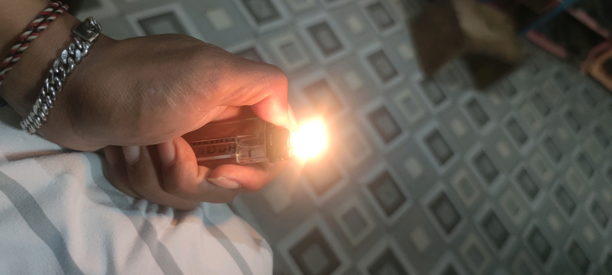 A person's hand, adorned with a silver chain bracelet and a red, white, and black woven bracelet, holds a lit, clear plastic lighter. The flame emits a bright orange-yellow glow. The background features a tiled floor with a pattern of interlocking squares in varying shades of gray. The image appears to be taken indoors, possibly in a residential setting in Gambarsari, Indonesia.