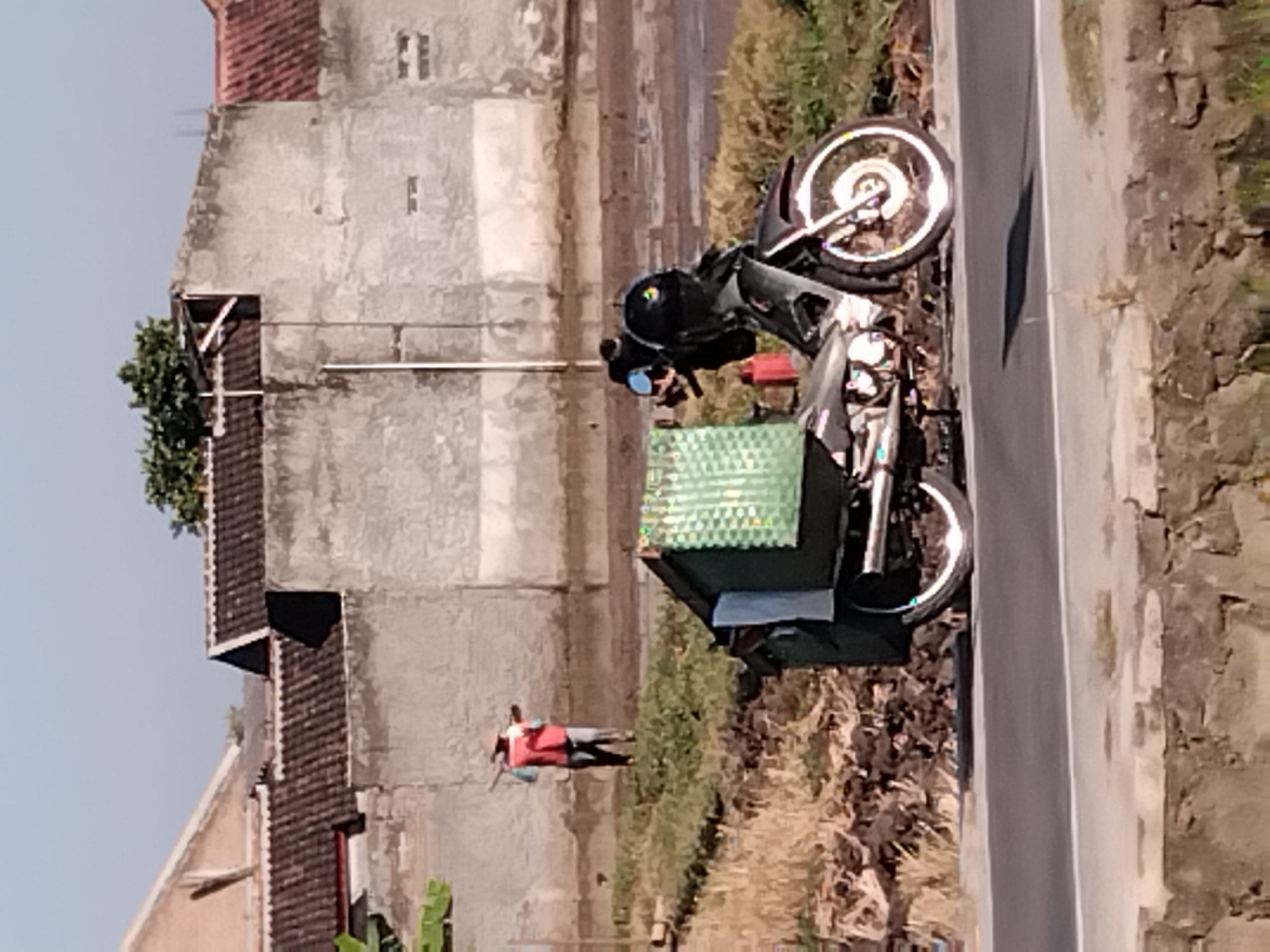 A motorcycle is parked on the side of a paved road in Papahan, Indonesia. The motorcycle is dark in color and has a large green box attached to its rear. Two helmets are placed on the motorcycle's handlebars. In the background, a person wearing a red vest and a hat is standing on a grassy embankment, holding a long object over their shoulder. Behind the person is a concrete wall with several buildings visible above it. The sky is clear and blue.