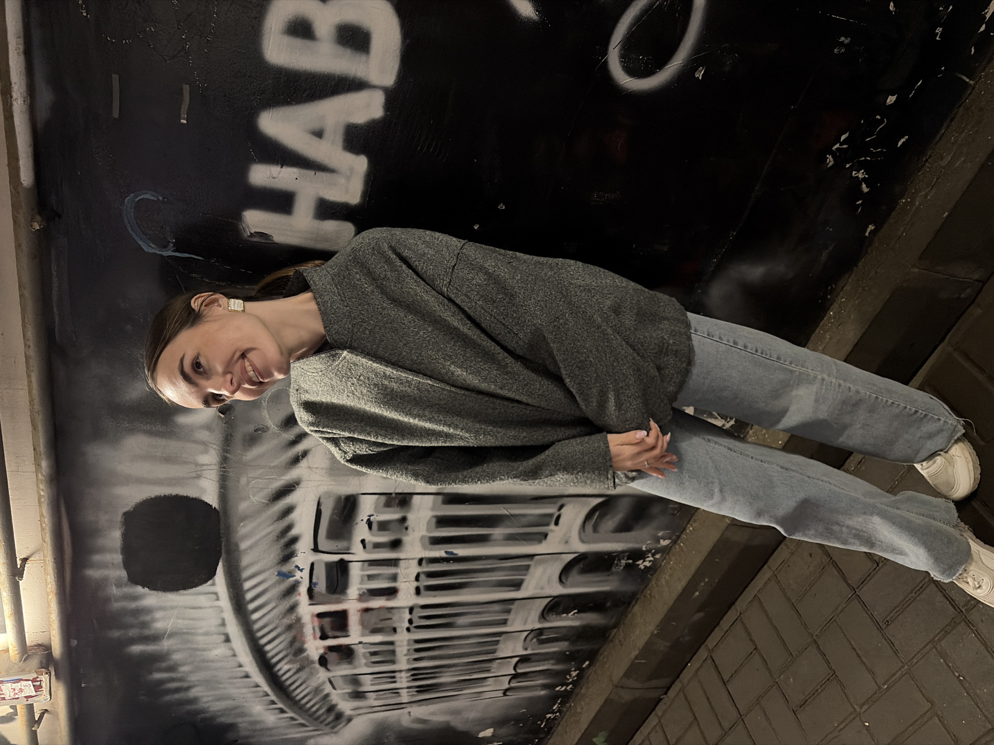 A young woman stands indoors against a graffiti-covered wall. She is wearing a gray sweater, light blue jeans, and white sneakers. The graffiti includes the word "HAB" and a drawing resembling a car grill. The scene appears to be in a pedestrian underpass, with brick pavement visible on the ground. The city or location is not apparent from this image.