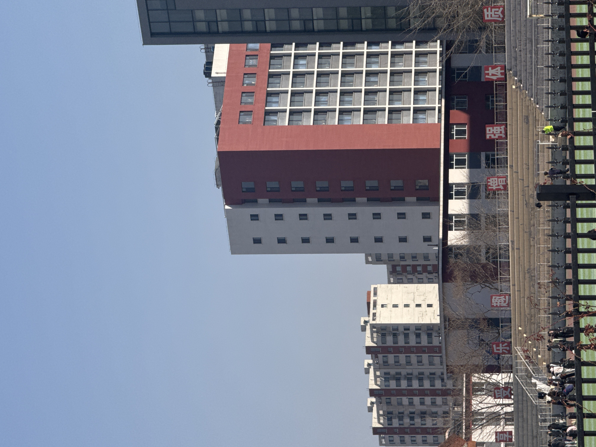 The image shows a building complex in Zhongtun, China. The buildings are predominantly white and red, with a grid-like window pattern. A covered walkway runs along the bottom of the frame, with signs in Chinese characters visible on the buildings behind it. People are walking along the walkway. The sky is clear and blue.