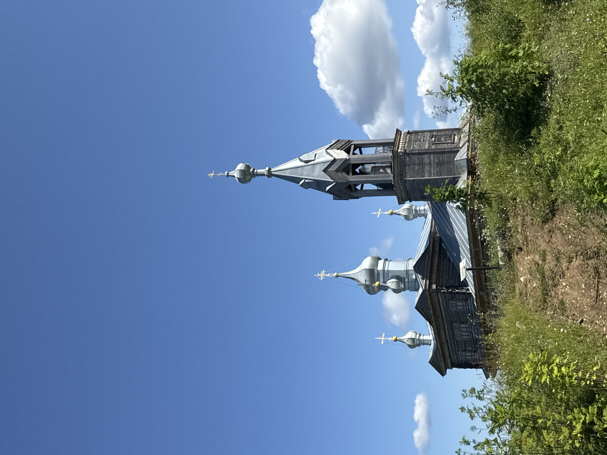 The image depicts an exterior view of the Church of St. Sergius of Radonezh in Cherdyn, Russia. The wooden church, with its several silver domes topped with crosses, is situated on a grassy hillside under a clear blue sky with scattered white clouds. The church features a prominent bell tower with an octagonal spire and open belfry. Lush green vegetation and wildflowers surround the church, with a dirt path leading up to it from the foreground. The scene captures a bright, sunny day, highlighting the architectural details of the historic Russian Orthodox church.