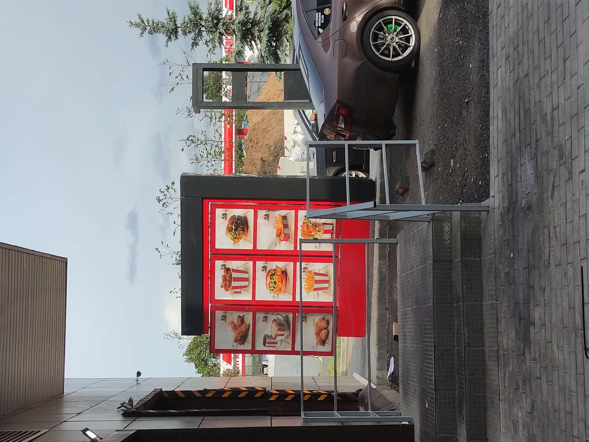 Here's a description of the image:
**Location:** Appears to be a drive-through at a fast-food restaurant. The city location is unknown.
**Subjects/Objects:**
* **Fast-food menu board:** Prominently displayed with images of food items (burgers, fried chicken, fries, etc.) in a red frame.
* **Car:** Part of a brown car is visible at the top right of the frame.
* **Metal barriers:** Rectangular metal barriers are in the foreground.
* **Building:** A building facade is visible on the left.
* **Sky:** The sky is cloudy.
* **Trees:** Scattered trees and landscaping are visible.
* **Drive-through speaker post:** There is a gray drive-through speaker box behind the metal barriers
**Scene Details:**
The image is taken at what appears to be a fast-food restaurant drive-through. A car is partially visible, presumably at the order station. The menu board is clearly visible, showing pictures of the available food. The scene has a somewhat urban, outdoor setting.