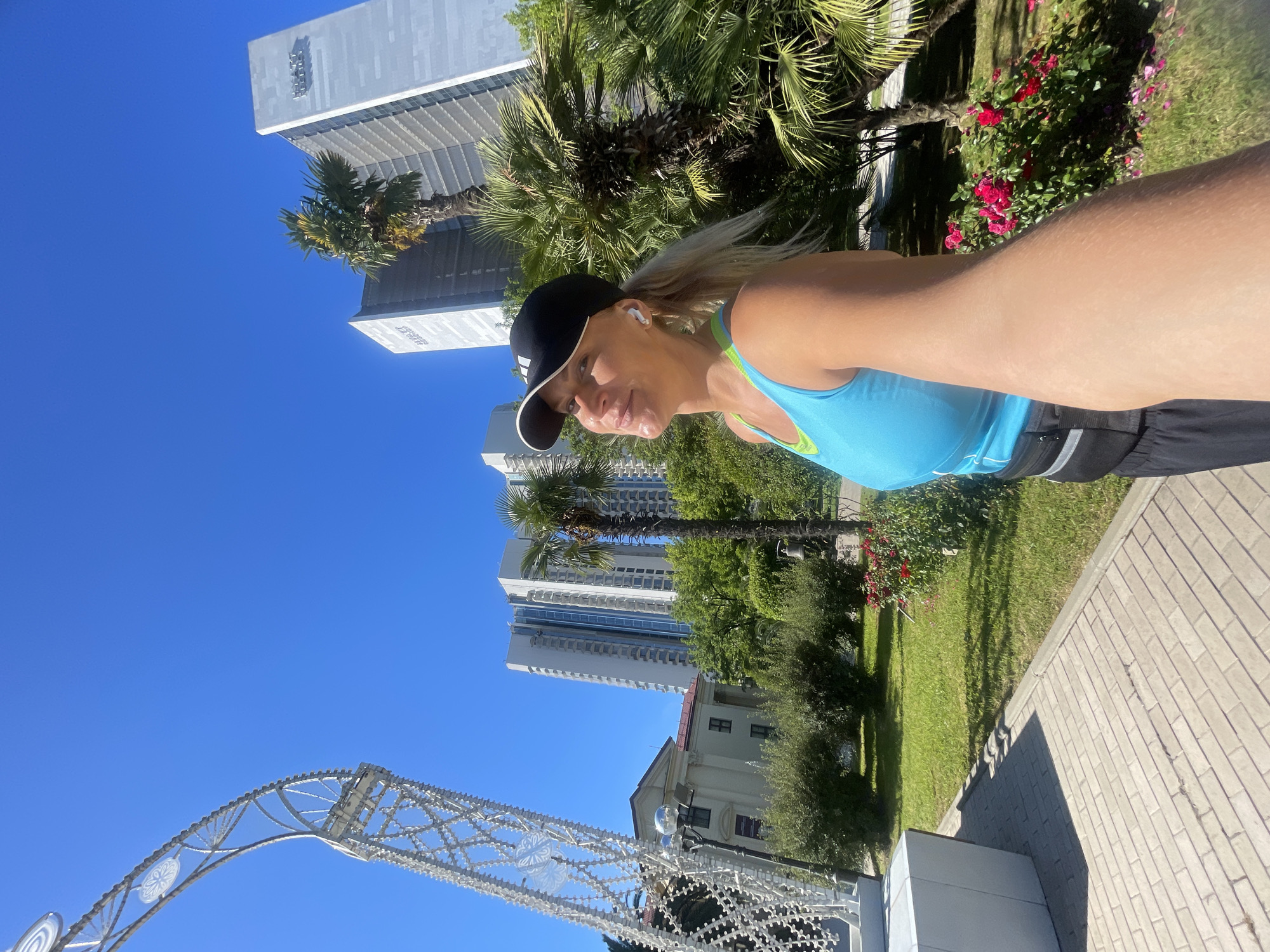 A blonde woman wearing a blue tank top, black athletic shorts, and a black baseball cap is taking a selfie outdoors in a city park. She has on white wireless earbuds and a black fanny pack. The woman is smiling as she looks up towards the sky. She is positioned in the foreground of the image, with the background showing a bright blue sky, several tall buildings, palm trees, green grass, and a paved walkway. The buildings appear to be modern high-rises, with one bearing the Hyatt logo. This scene is located in the city of Sochi, Russia, likely in a resort area given the presence of palm trees and modern architecture. The sun is shining brightly, casting shadows on the ground. To the left of the woman, a large, white, ornate arched structure is visible. Red roses are blooming in bushes behind her.