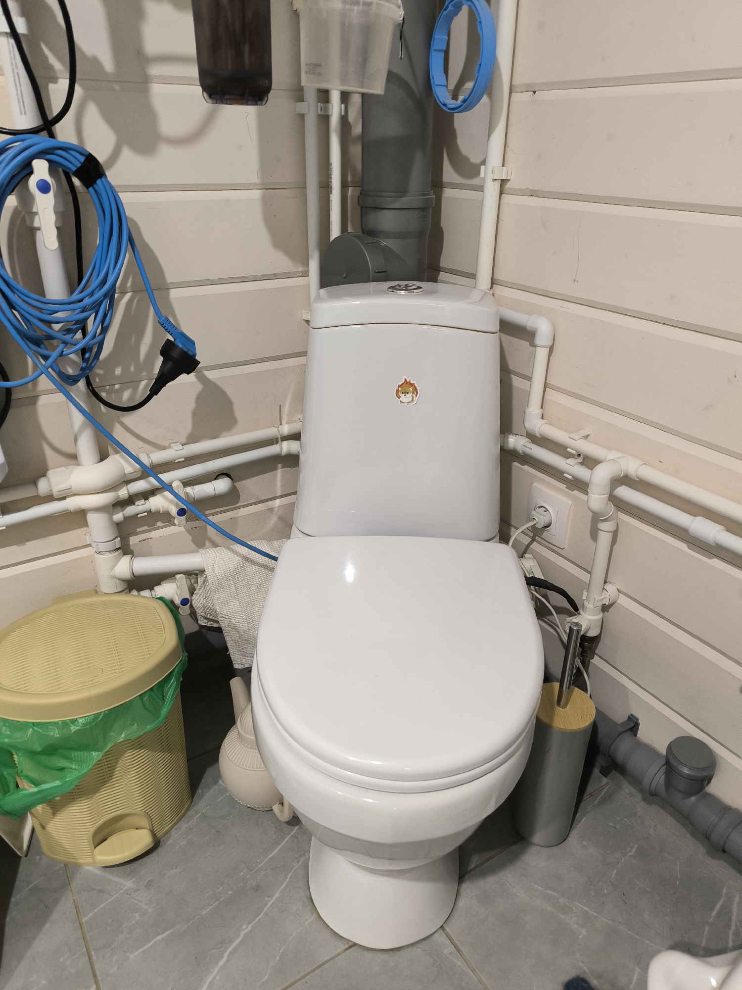 The image shows a white toilet in a bathroom. There is a yellow trash can with a green liner to the left of the toilet. A toilet brush holder is to the right of the toilet. The walls are paneled with light-colored wood. Plumbing pipes are visible throughout the scene. A blue electrical cord is coiled on the wall to the left. This scene is consistent with a residential bathroom in Октябрьский, Russia.