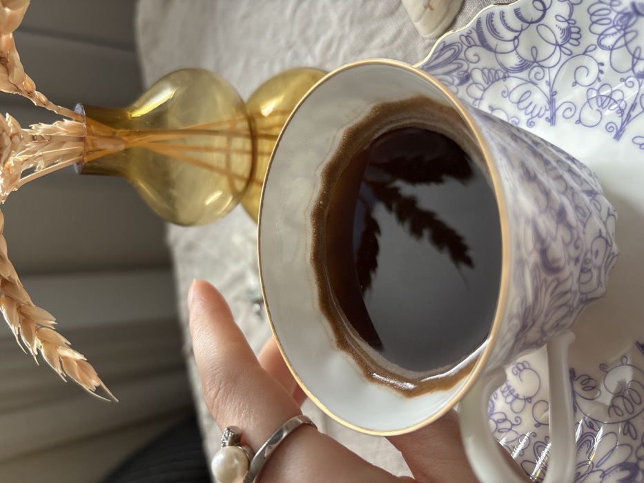 An indoor, close-up image shows a person's right hand holding a white porcelain cup filled with a dark liquid, likely coffee, topped with a thin layer of crema. The cup features a gold rim and intricate purple scrollwork patterns, resting on a matching saucer. A silver-colored ring, adorned with a white pearl and small clear stones, is visible on the ring finger of the hand. In the background, an amber-colored glass vase containing several dried wheat stalks stands on a light-colored, textured surface, possibly a tablecloth. The shadow of the wheat stalks is reflected in the liquid within the cup. A white seashell is partially visible on the right side of the frame. This scene is located in the city of Kudrovo, Russia.