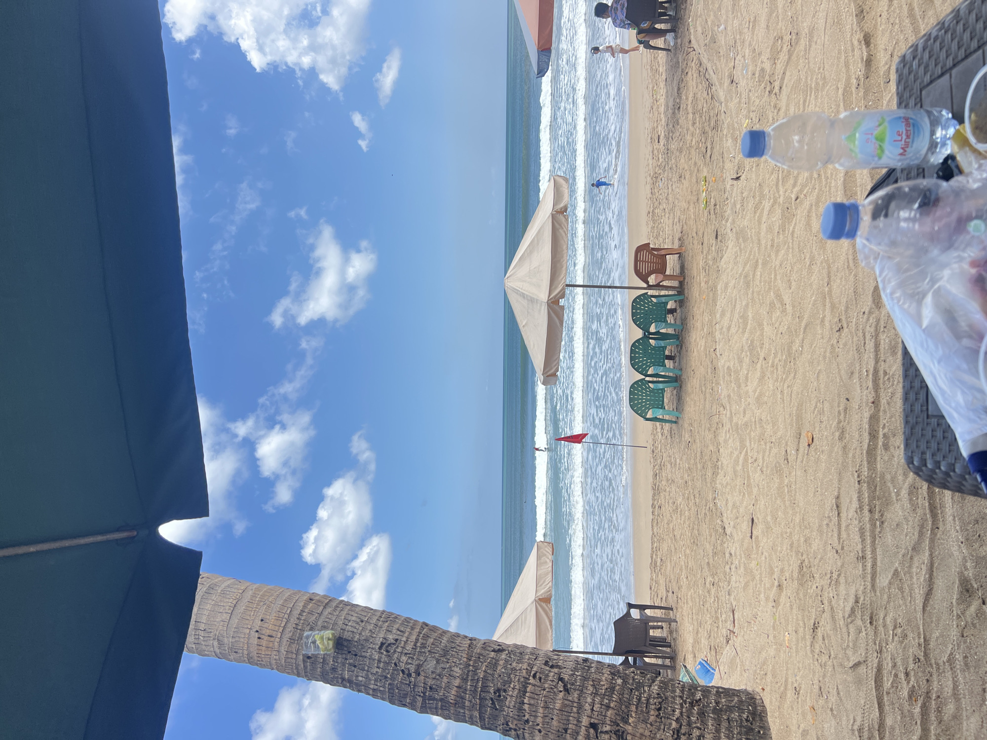 The image depicts a beach scene in Kuta, Indonesia, under a bright blue sky with scattered white clouds. In the foreground, two plastic water bottles are on a dark-colored table. One bottle, labeled "Le Mineral," is partially filled with water and ice. A plastic bag is also visible on the table.
The middle ground of the image shows a sandy beach with several plastic chairs arranged in groups. Two beige beach umbrellas are set up, one more prominent than the other. A red flag is planted in the sand near the water's edge. The ocean is visible with moderate waves breaking on the shore. A surfer is present in the water, and a young child is wading near the shore.
In the background, beyond the beach, the ocean stretches towards the horizon. A tall palm tree with rough bark is positioned to the left of the frame, partially obscuring the view of the sky and ocean. A small glass jar containing what appears to be lime slices is attached to the palm tree. Two individuals are seated on chairs further down the beach, with their backs to the camera.