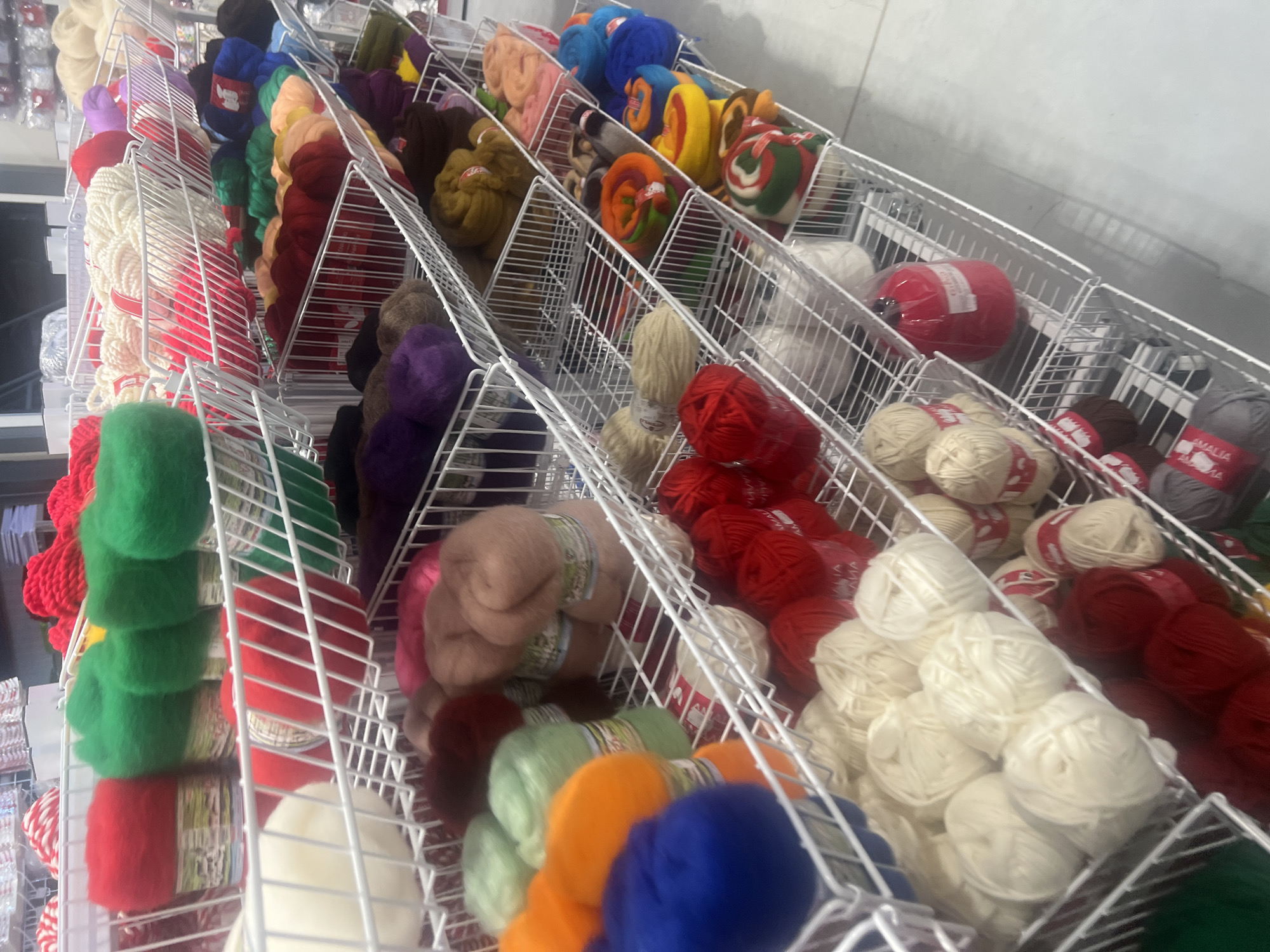 The image shows shelves of yarn in a retail setting, likely a yarn or craft store. The shelves are made of white wire mesh, and are filled with various colors and types of yarn. The foreground features green, red, and white yarn, followed by a row of diverse colors including orange, blue, green, purple, and brown. In the background, there are more colorful skeins and spools of yarn. The location is not directly identifiable from the image, but the presence of "Amalia" brand yarn suggests it may be in Burgas, Bulgaria.