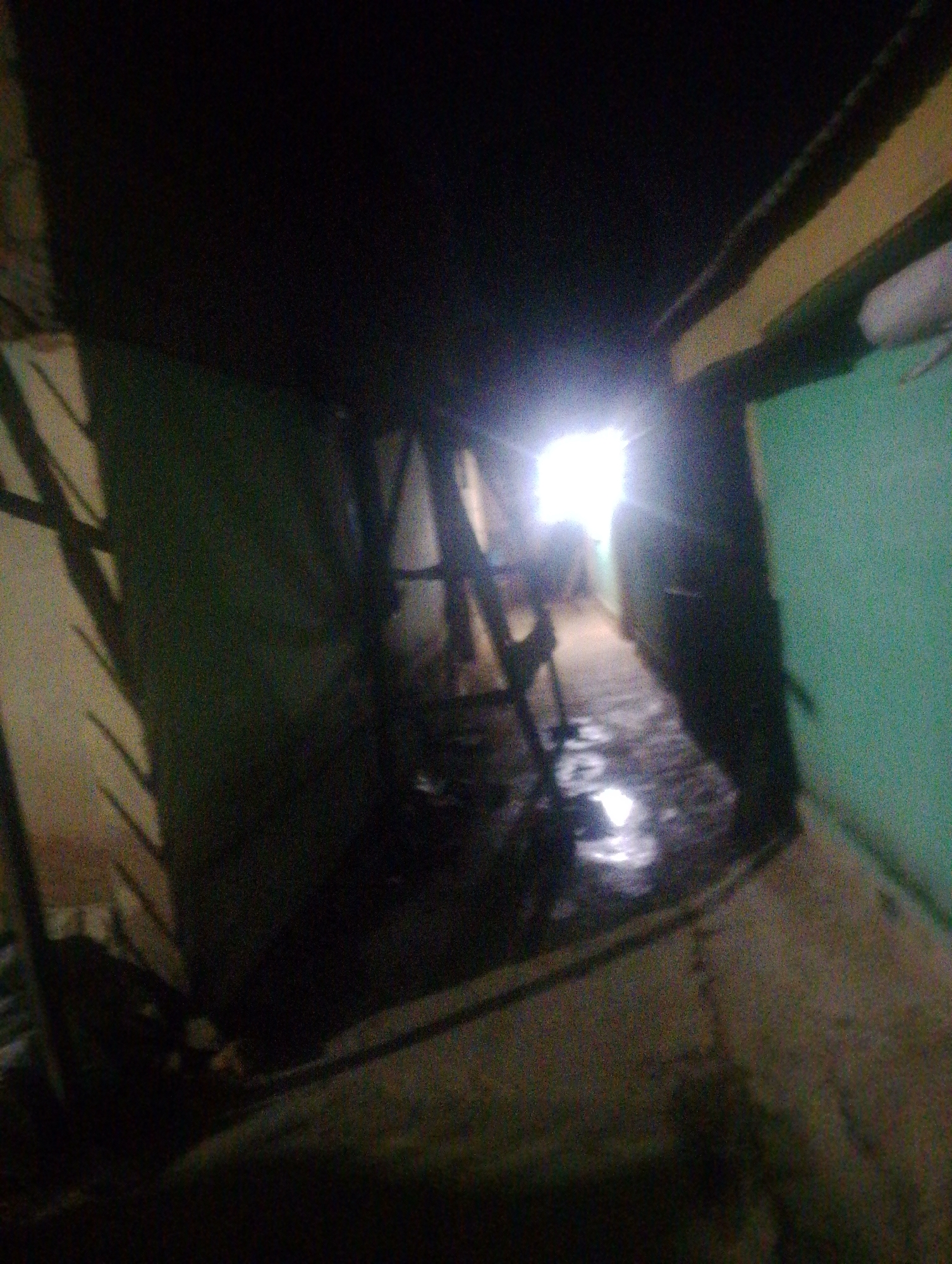 The image depicts the interior of a dimly lit, narrow passageway or corridor. A bright light source emanates from the far end of the corridor, suggesting an opening such as a door or window. The walls appear to be painted in muted colors, with a green section visible on the right side. The floor is wet and reflective, indicating the presence of water or a similar liquid. Structural elements like beams and what appear to be diagonal supports are visible along the ceiling and walls, suggesting a utilitarian or possibly unfinished structure. There is debris or dark material scattered on the wet floor in the middle ground, and some unclear objects are present in the shadows. The overall impression is one of a neglected or abandoned space. The location is identified as Jos, Nigeria, although specific details within the image do not confirm this location beyond the provided context.