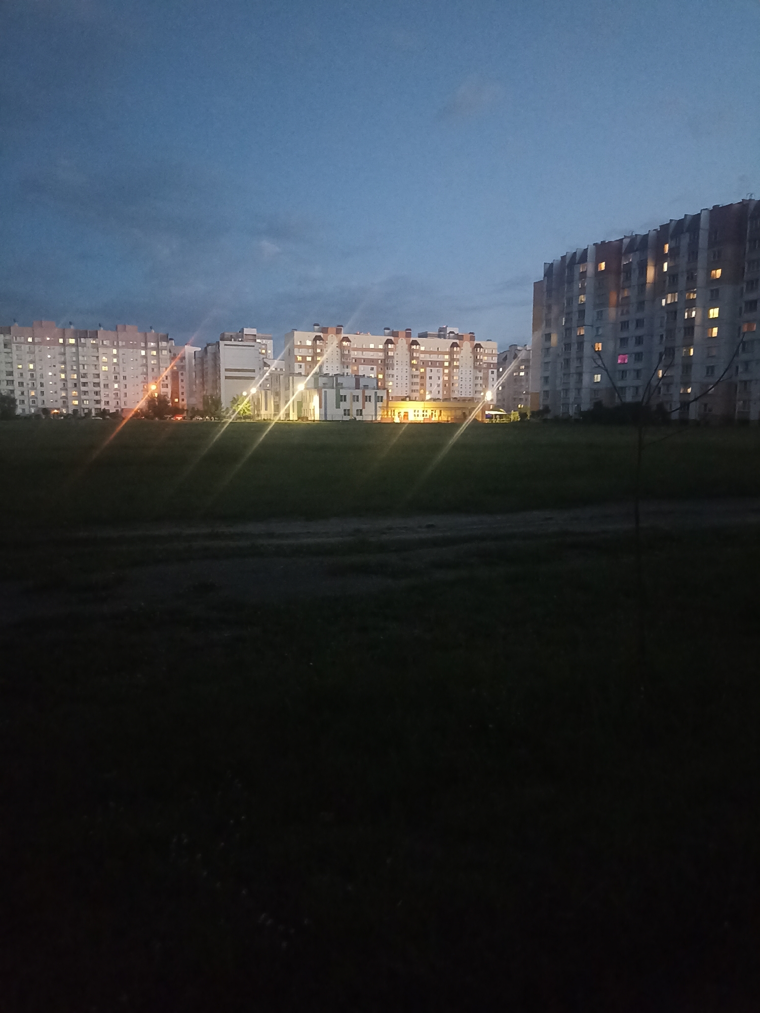 In the city of Homyel, Belarus, at dusk, multiple residential buildings stand illuminated by interior lights, casting a warm glow against the darkening sky. The buildings are of varying heights and architectural styles, with some featuring balconies and pitched roofs. A vast expanse of green grass stretches out in front of the buildings, a stark contrast to the urban landscape. The image is characterized by an eerie stillness, with no visible people or signs of activity, hinting at a quiet evening in this Belarusian city.