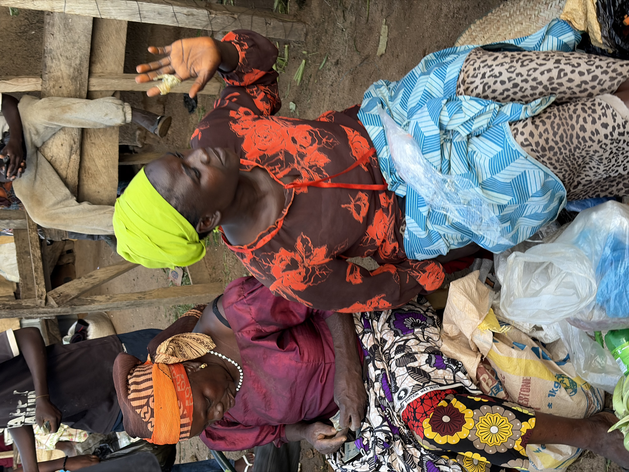 In Gura Top, Nigeria, two women are seated at what appears to be a market stall. The woman on the left, wearing a burgundy top and a colorful wrap skirt, is counting money. She has a pearl necklace and an orange and brown headwrap. The woman on the right, in a brown shirt with orange floral patterns and a bright green headscarf, gestures with her right hand, showing a band-aid on one finger. She is also wearing a blue and white striped wrap. Several sacks and plastic bags are visible around them, suggesting they are vendors or shoppers. Wooden structures, possibly part of the market, are in the background. The ground is dirt.