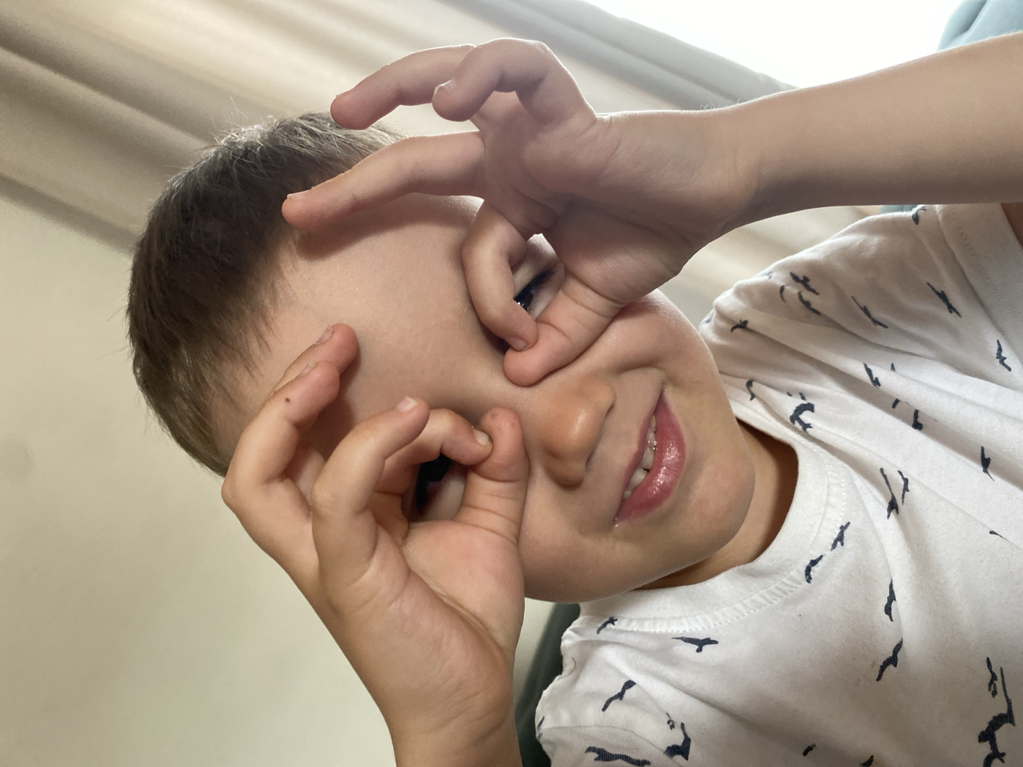 A close-up, slightly angled, indoor shot shows a young boy making circles with his fingers around his eyes, appearing to mimic eyeglasses. The boy has short, dark brown hair and fair skin. He's wearing a white T-shirt with a pattern of small, dark blue birds. The background is a plain, light beige wall. There is no information in the picture to confirm the location in Chaikovsky, Russia.