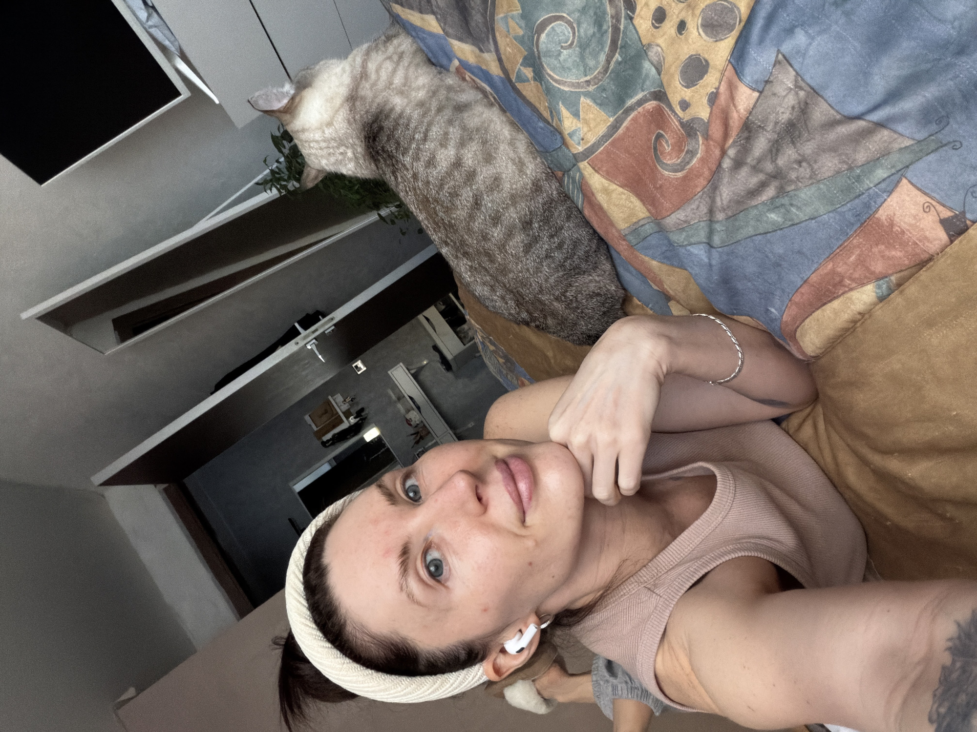 The media file features a selfie taken by a woman indoors, with her cat nearby. The woman, in the foreground, is lying down and looking directly at the camera with wide, light-colored eyes and a relaxed, slightly smiling expression. She has fair skin, dark hair pulled back, and is wearing a cream-colored braided headband and a light pink ribbed tank top. A white earbud is visible in her right ear, and a silver chain bracelet adorns her left wrist. A tattoo is partially visible on her left arm.
Beside her, a grayish-brown domestic cat, likely a tabby or spotted breed, is seen from the back and side, lying on a colorful patterned blanket and looking away towards the background. The blanket features abstract and geometric designs in shades of blue, yellow, orange, and brown. Beneath it, a plain brown or tan blanket is visible.
The background shows an indoor room, possibly a bedroom or living area, with light-colored shelves or ledges. A small green plant is partially visible behind the cat. The lighting appears to be ambient indoor light. The scene depicts a casual and serene moment at home in Perm, Russia. No visible text is present in the image.