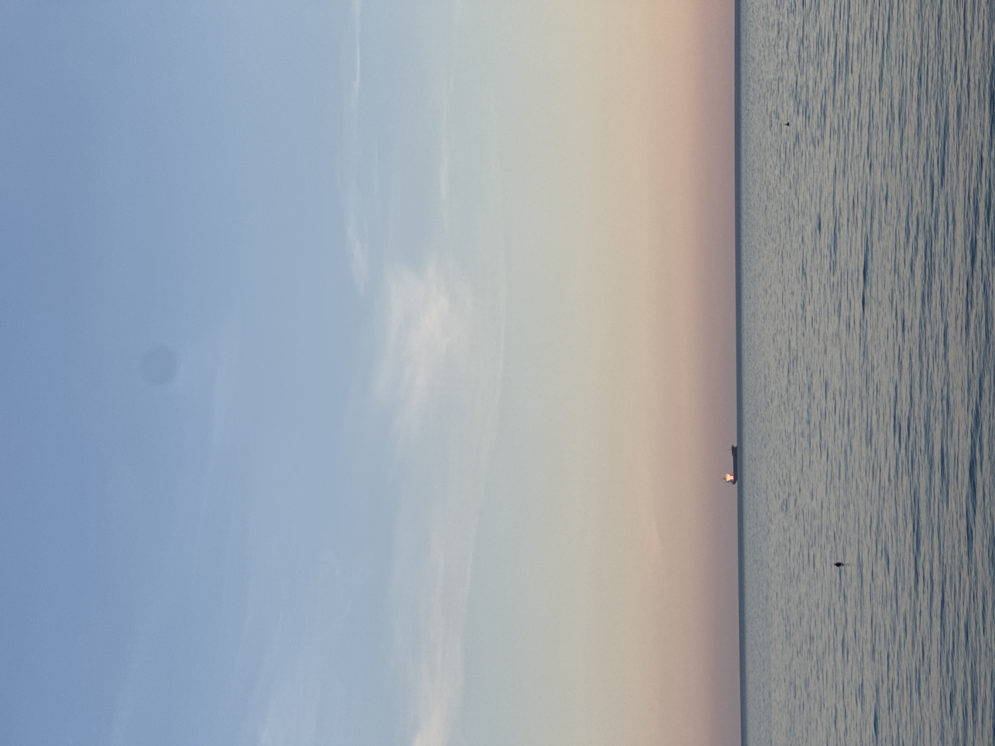 The image depicts a seascape under a sky gradating from blue to pale orange, likely at dawn or dusk. The right side of the frame is occupied by the sea, showing subtle wave patterns. On the horizon, there is a faint suggestion of land. A single object, possibly a small boat or buoy, is visible near the horizon line. The sky dominates the left half of the image, with a smooth transition in color. This location is likely in Sutomore, Montenegro, given the coastal environment.