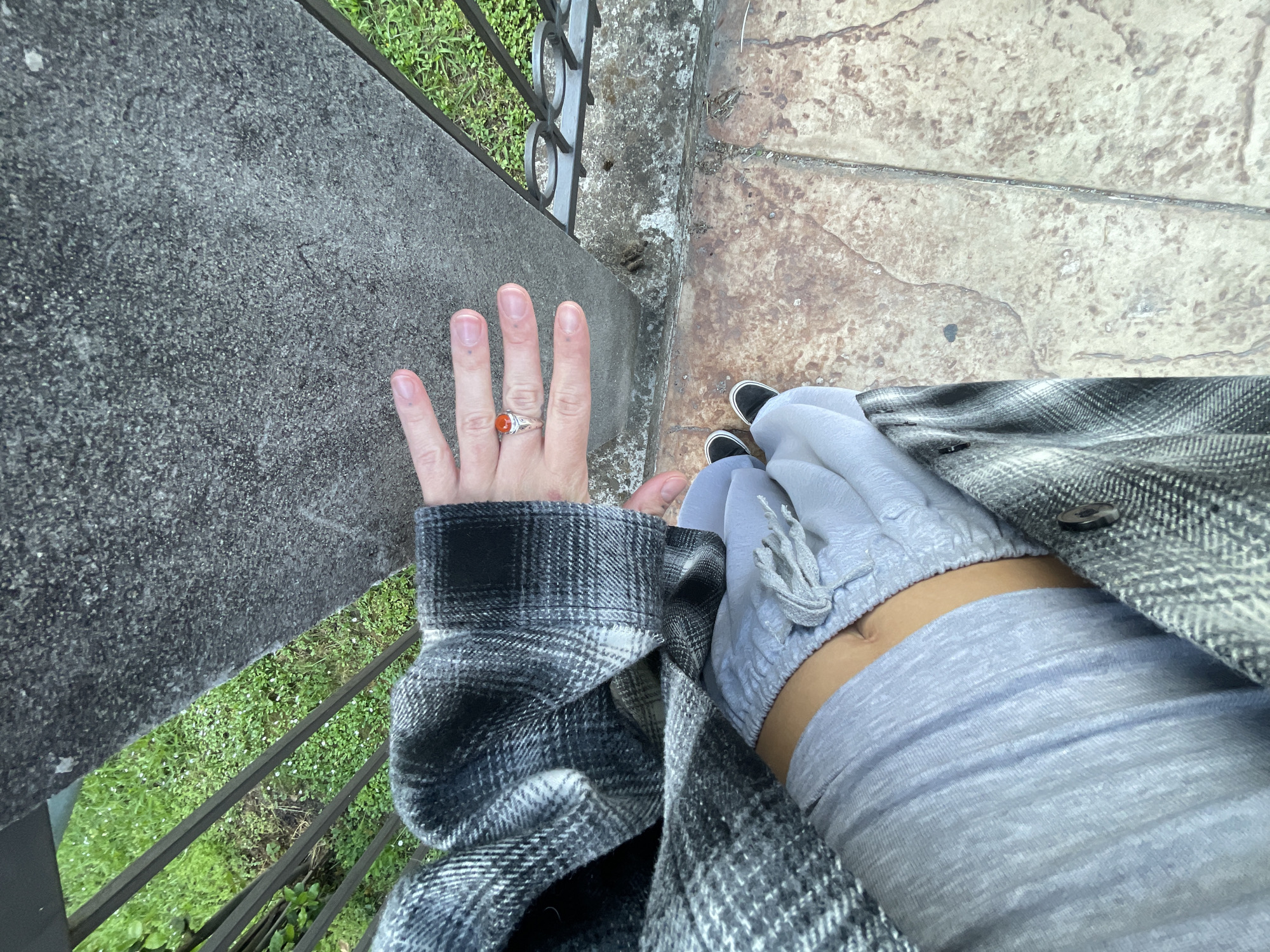 A first-person, high-angle shot depicts a person standing on a paved surface adjacent to a weathered concrete barrier with a metal fence along its edge. The person wears a gray and black plaid jacket over a light blue long-sleeved shirt. The jacket sleeve is pushed up to reveal the person's wrist and hand, adorned with a silver ring featuring a prominent orange stone on the ring finger. The hand is extended flat against the concrete barrier.
The paved surface transitions into a tan tile or stone pattern. The person's feet, clad in black canvas sneakers, are visible near the tile surface.
The scene hints at an outdoor location near vegetation, as glimpses of green grass are visible through the metal fence. Based on the architectural style and the specific combination of paving, concrete barriers, and wrought iron fencing, the location is likely Budva, Montenegro. The image captures a moment of observation, with the person seemingly pausing to rest their hand on the barrier while standing on the walkway.