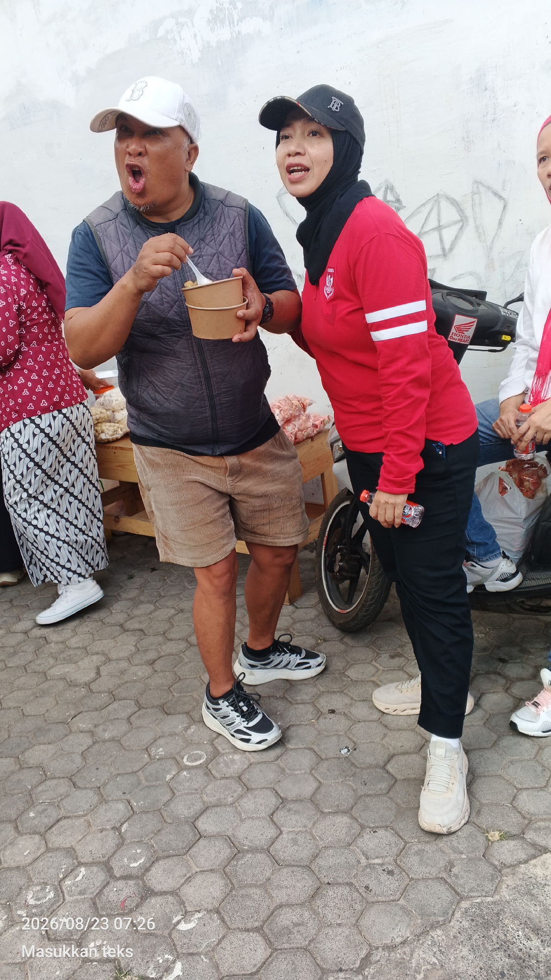 In Malang, Indonesia, on a paved ground surface composed of hexagonal gray interlocking stones, a group of people are gathered. In the foreground, a man wearing a white cap, a grey vest over a dark shirt, and tan corduroy shorts is holding two brown paper bowls and a plastic spoon, with his mouth open as if surprised or exclaiming. To his right, a woman in a black hijab and a red long-sleeved shirt with white stripes on the sleeves is looking towards the man. She is holding a small plastic bottle with a red cap in her right hand. Behind them, a wooden table is laden with bags of what appears to be snacks. To the left of the man, a person wearing a maroon patterned top and a white and black patterned skirt is partially visible. The background features a white wall with some charcoal drawings. A motorcycle is parked to the right of the woman. In the bottom left corner of the image, a timestamp reads "2026/08/23 07:26".