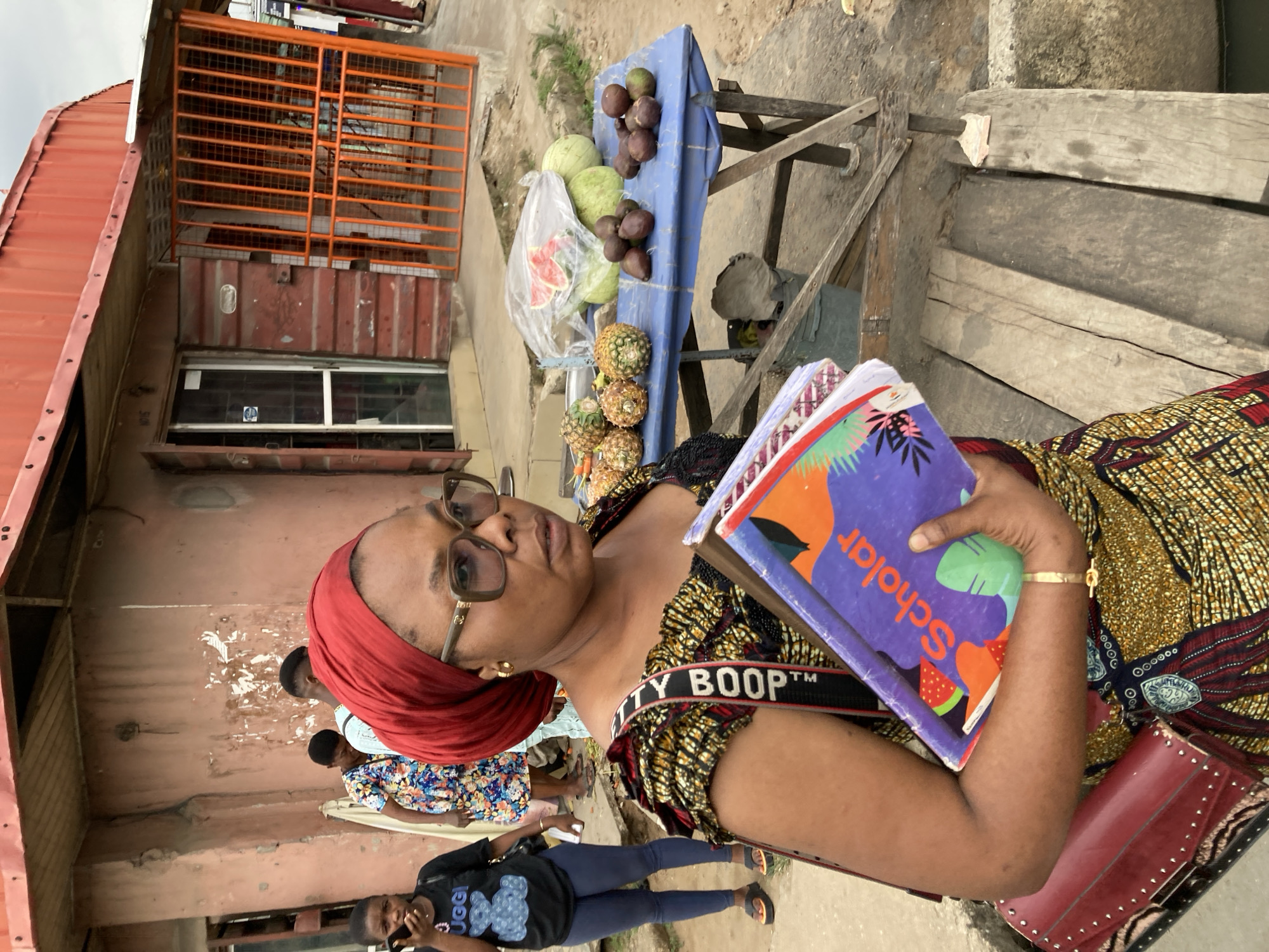 A woman with a red headwrap and glasses stands beside a fruit stand on a wooden table. The stand holds pineapples, watermelons in a plastic bag, and purple fruits on a blue cloth. The woman holds a stack of books with the word "Scholar" visible on the cover. She wears a yellow and brown patterned dress with a "Betty Boop" strap across her shoulder, and carries a red purse. In the background, a shipping container converted into a building has an orange metal gate and a window. Behind the woman, other people walk along the roadside. The location is likely in Aba, Nigeria, based on the style of the building and the items on sale.