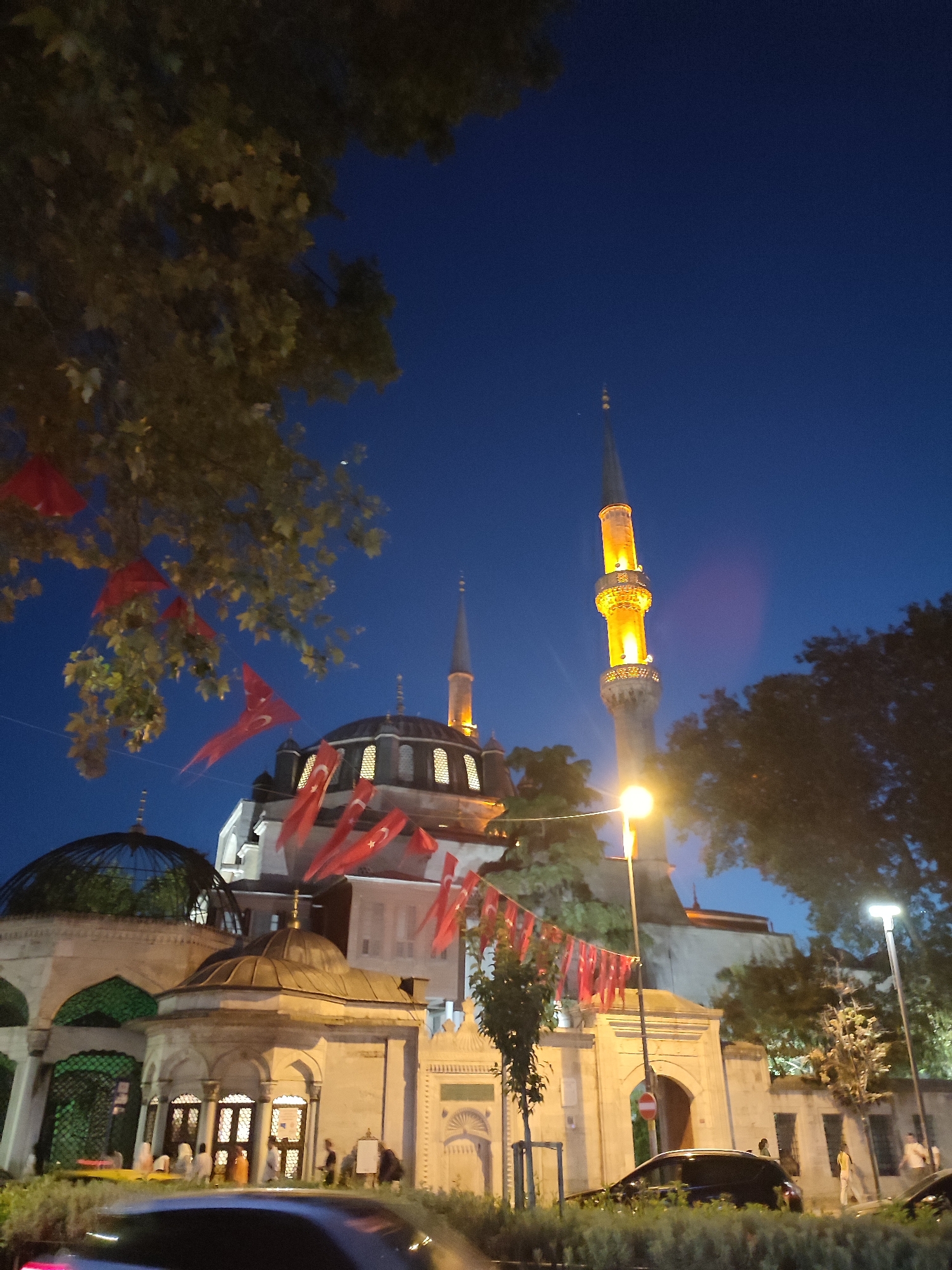 The image shows the Mihrimah Sultan Mosque and its complex in Üsküdar, Turkey, at dusk. The mosque, a prominent Ottoman-era structure, is illuminated, highlighting its domes and minarets. Red flags with crescent moons and stars, likely Turkish flags, are strung across the scene, adorning trees and fences. Several people are visible near the entrance of one of the buildings within the complex, some appearing to be walking or gathered. Cars are parked or driving on the street in the foreground, and streetlights provide additional illumination. The sky is a deep blue, indicating twilight.