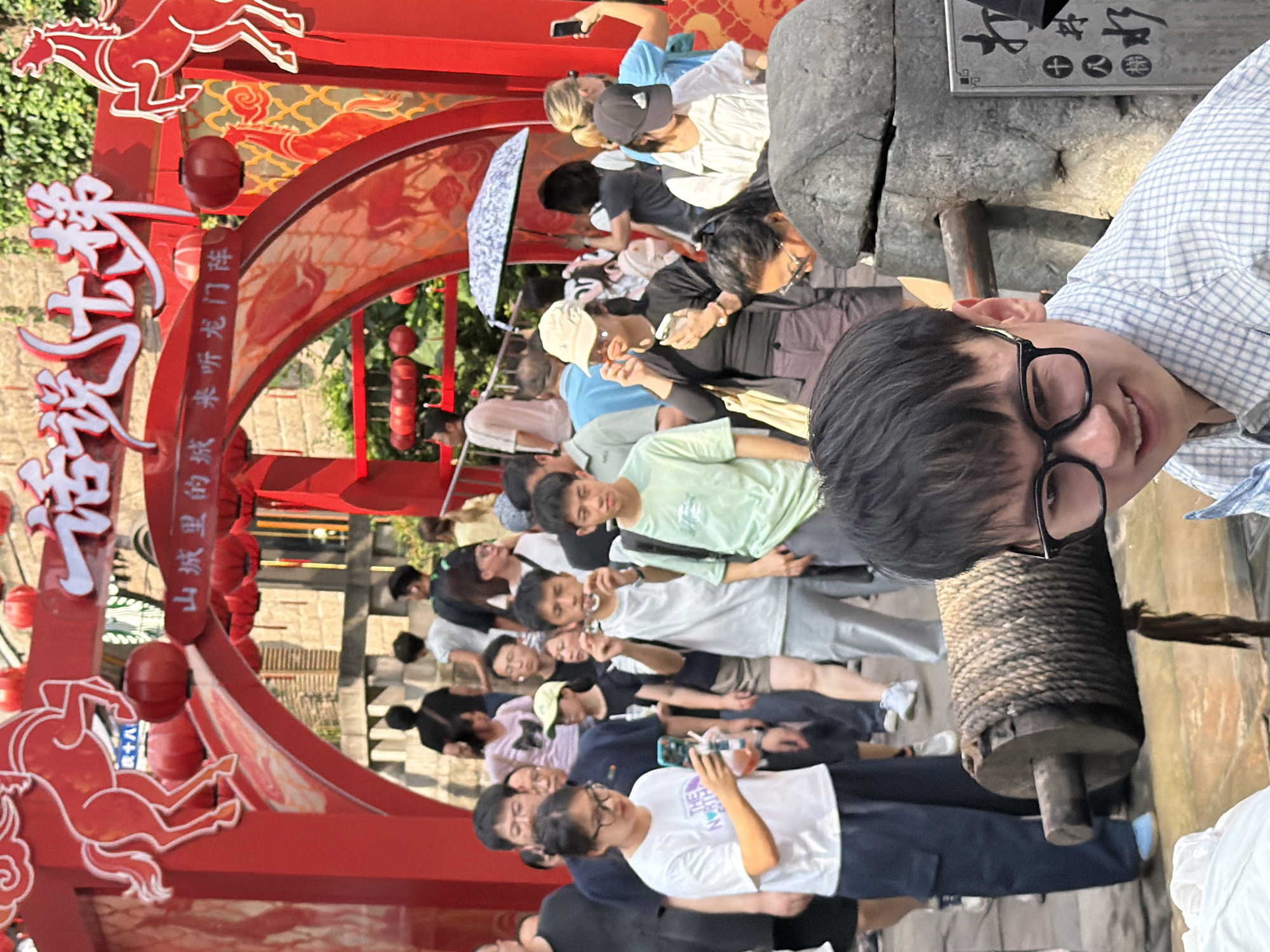 In Yuzhong District, China, a group of people are gathered near a decorative red archway adorned with images of horses and red lanterns. A young man with glasses in the foreground is smiling, facing the camera. Behind him, other individuals are looking at their phones, holding umbrellas, and walking. The archway features Chinese calligraphy. The scene appears to be an outdoor market or tourist attraction.