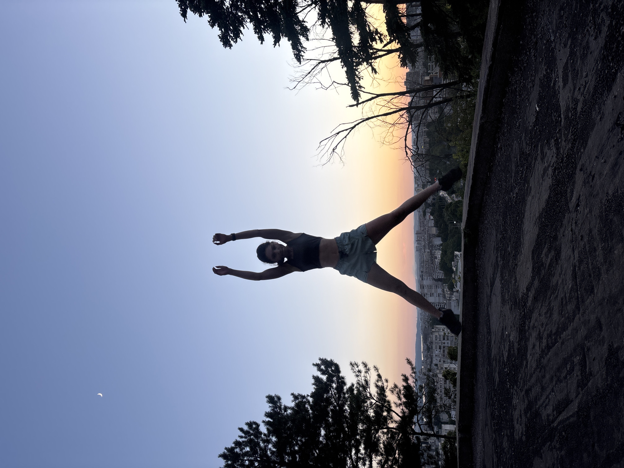 A silhouette of a woman in athletic wear is captured mid-jump against a twilight sky over Varna, Bulgaria. The woman's arms are raised overhead, and her legs are spread wide. She is positioned on a concrete ledge, overlooking a cityscape with buildings and trees. A crescent moon is visible in the upper left corner of the sky, and a gradient of orange and pink hues suggests the setting sun. The foreground is a dark, textured concrete surface.