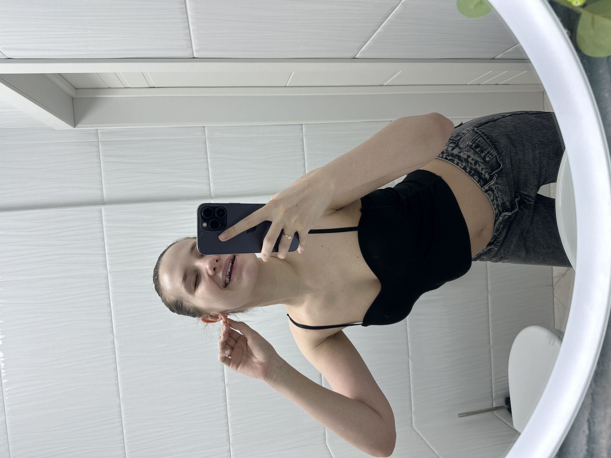 A young adult female is depicted in a mirror selfie taken within a modern bathroom. While stated to be in Moscow, Russia, no specific identifying markers for the city are visually present in the image. The subject, with dark hair pulled back, is smiling broadly with eyes partially closed, revealing dental braces on her upper teeth. She wears a black spaghetti-strap crop top and dark grey, washed-out jeans. Her left hand holds a dark-colored smartphone, featuring a multi-lens camera array, used to capture the image; a metallic ring adorns her left ring finger. Her right hand is positioned at her right ear, appearing to adjust a small, metallic ear accessory. The bathroom walls are clad in white, rectangular tiles exhibiting a subtle wavy texture. A white, modern door with horizontal paneling is visible in the reflection to the right. The lower left portion of the reflection shows part of a white floor-mounted toilet and a toilet brush in a holder beside it. The floor appears to be a light-colored, possibly tiled, surface. The mirror itself has a prominent white, oval-shaped frame, and a blurred green object, possibly a decorative plant, is reflected in the lower right corner.