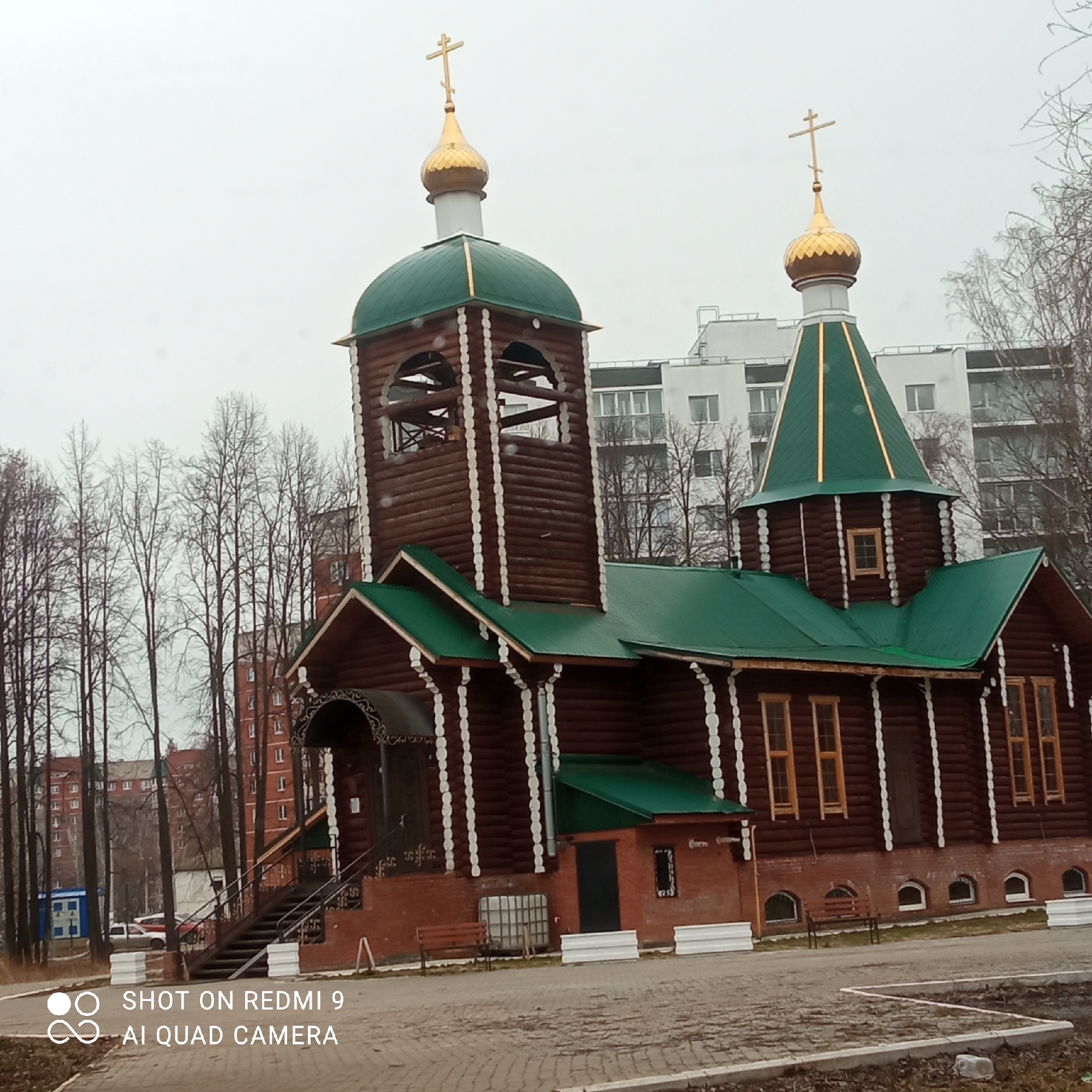 The image captures a wooden Russian Orthodox church under an overcast, dull sky, likely in Chaikovsky, Russia. The church is constructed from dark brown logs, accented by distinct white vertical decorative trim, and features vibrant green metal roofs. Two golden onion domes, each topped with a golden Orthodox cross, stand out against the light sky. The larger dome crowns a prominent bell tower, while the smaller one sits atop the main church structure.
The building rests on a brick foundation, where small arched windows indicate a basement level. The main entrance, located on the left side of the building, is accessed via dark stairs with metal railings and is covered by an ornate, dark overhang. A small, single-story brick extension with a green roof is attached to the side of the church, near which a large white plastic container is visible. Several brown benches are placed along the paved walkway surrounding the building.
The background features numerous tall, bare deciduous trees, suggesting a cold season such as late autumn, winter, or early spring. Beyond the trees, modern multi-story residential buildings are visible, indicating an urban or suburban setting. The foreground is paved with interlocking bricks. No people are present, and no specific activity is taking place. In the bottom left corner, the text "SHOT ON REDMI 9 AI QUAD CAMERA" is visible as a watermark.
