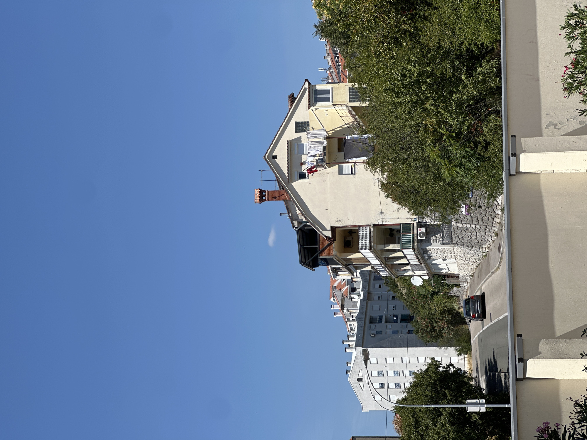 The image captures a street scene in the city of Rijeka, Croatia, under a clear blue sky. On the right side of the road, a pale yellow building with red-tiled roofs and multiple balconies is prominent. Laundry is drying on a clothesline attached to one of the balconies, and an air conditioning unit is visible. The building is partially obscured by lush green trees.
Across the street, a grey car is driving up a winding road. To the left of the road, a tall, multi-story grey building with numerous windows stands out. Further in the background, more buildings with red roofs can be seen, suggesting a dense urban environment. A street lamp with a white fixture is positioned on the left side of the road. In the foreground, a cream-colored wall with a grey trim runs horizontally across the bottom of the image, with some flowering plants visible on the left. The scene appears to be a residential area.