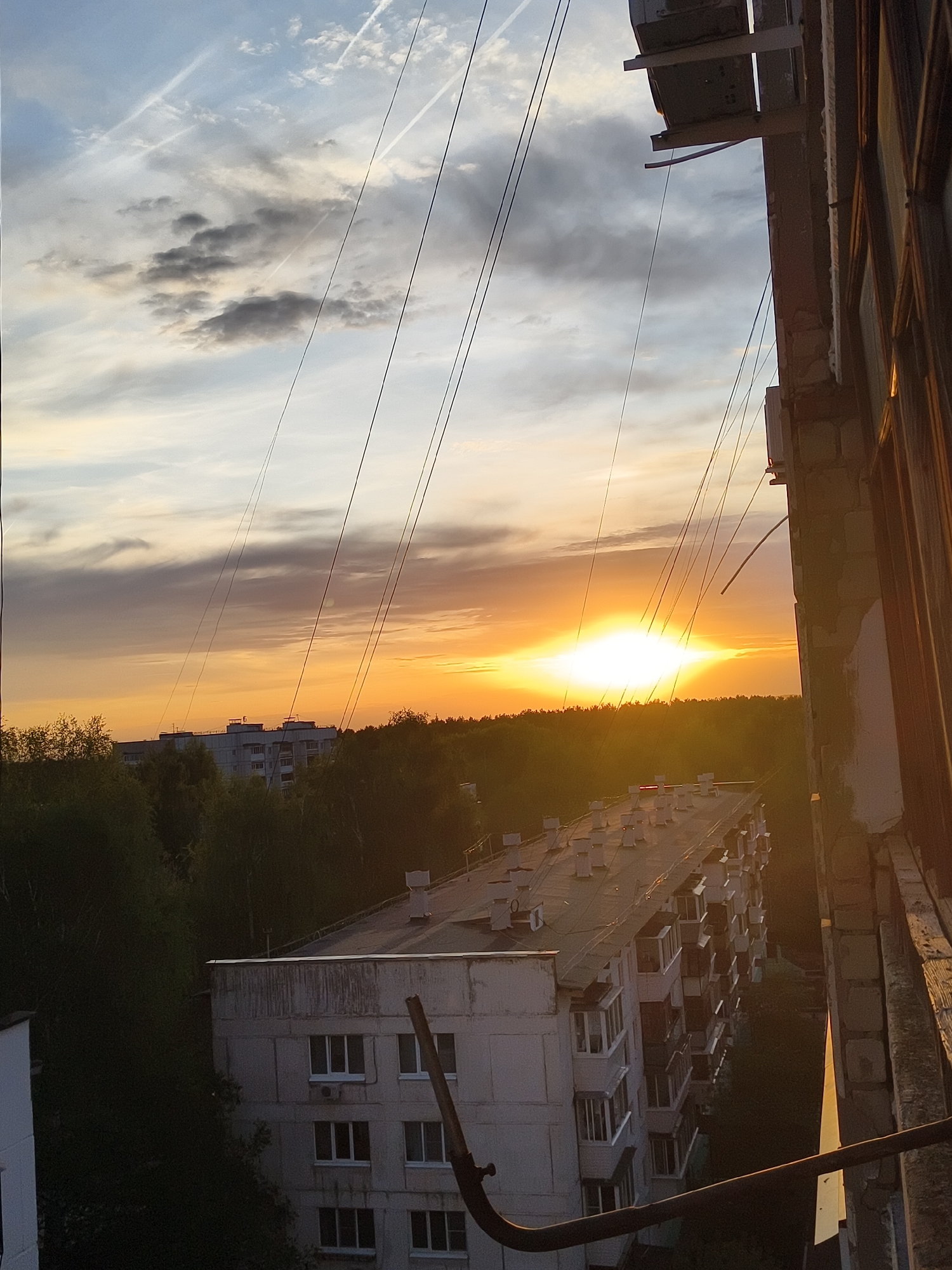 The image is a high-angle shot taken from an apartment balcony in the city of Unknown, Unknown. The time of day appears to be sunset, with a bright orange sun positioned near the horizon. The sky above features a mix of gray and light clouds, with visible contrails from aircraft.
In the foreground, the balcony structure is visible, including concrete walls and metal railings. Several wires stretch across the view towards the horizon, likely power or telephone lines.
Below, there are several residential buildings surrounded by dense green trees. The buildings are typically low to mid-rise, constructed from concrete, with flat rooftops that have multiple chimneys and air conditioning units. The architectural style of the buildings suggests that it is a residential area in a city, possibly in Eastern Europe or Russia.
No people are visible in the shot. The overall scene suggests a quiet, urban environment at dusk.