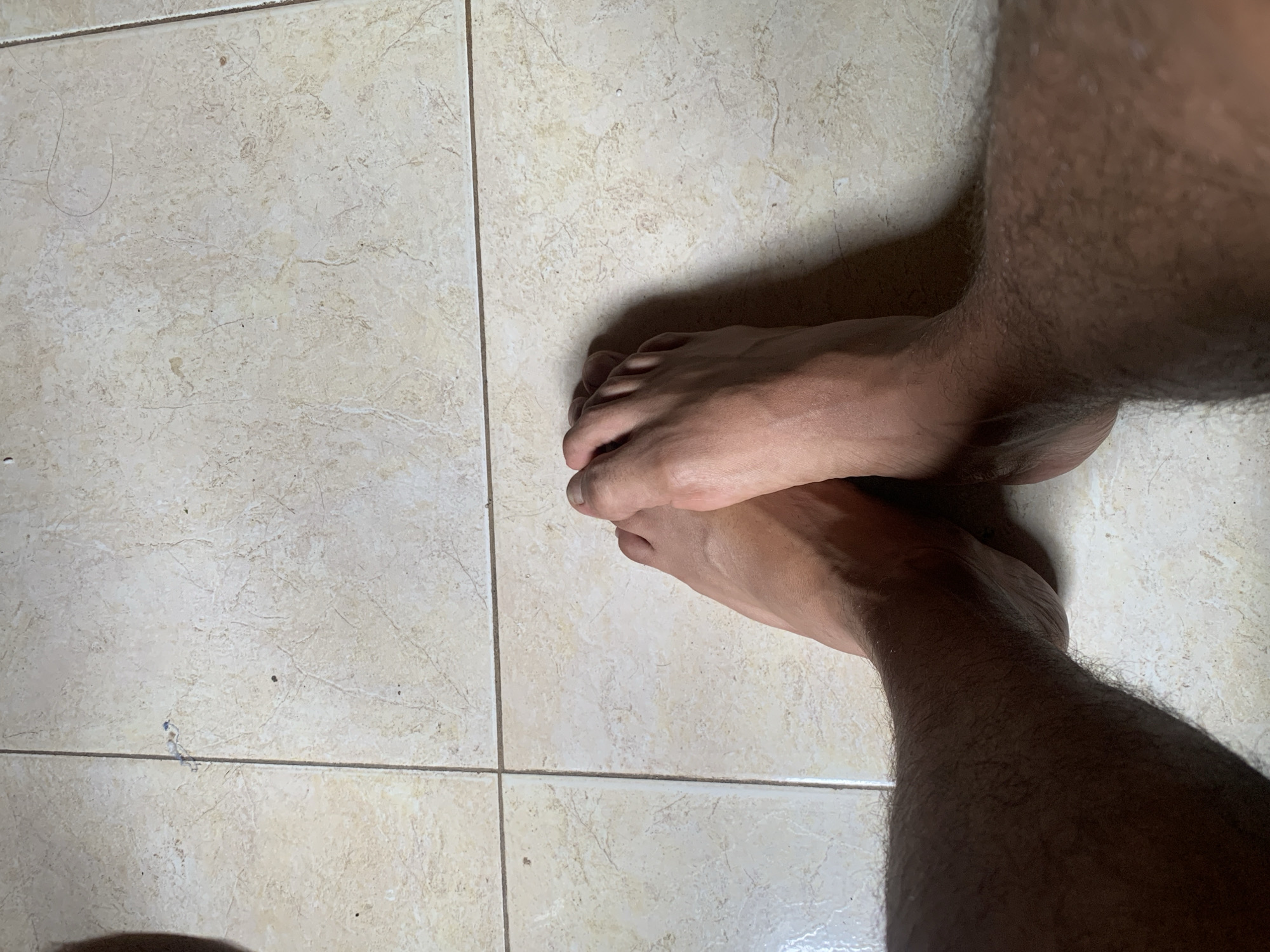 The image shows a close-up, high-angle shot of a pair of bare feet resting on a tiled floor. The feet are positioned with the toes of both feet touching, and the legs are visible from the knees down, covered in dark hair. The floor is composed of light-colored square tiles with a marbled texture and visible grout lines. The lighting suggests an indoor environment, likely a home, given the casual placement of the feet. The scene does not provide specific details to place it in Santa Ana, Venezuela.