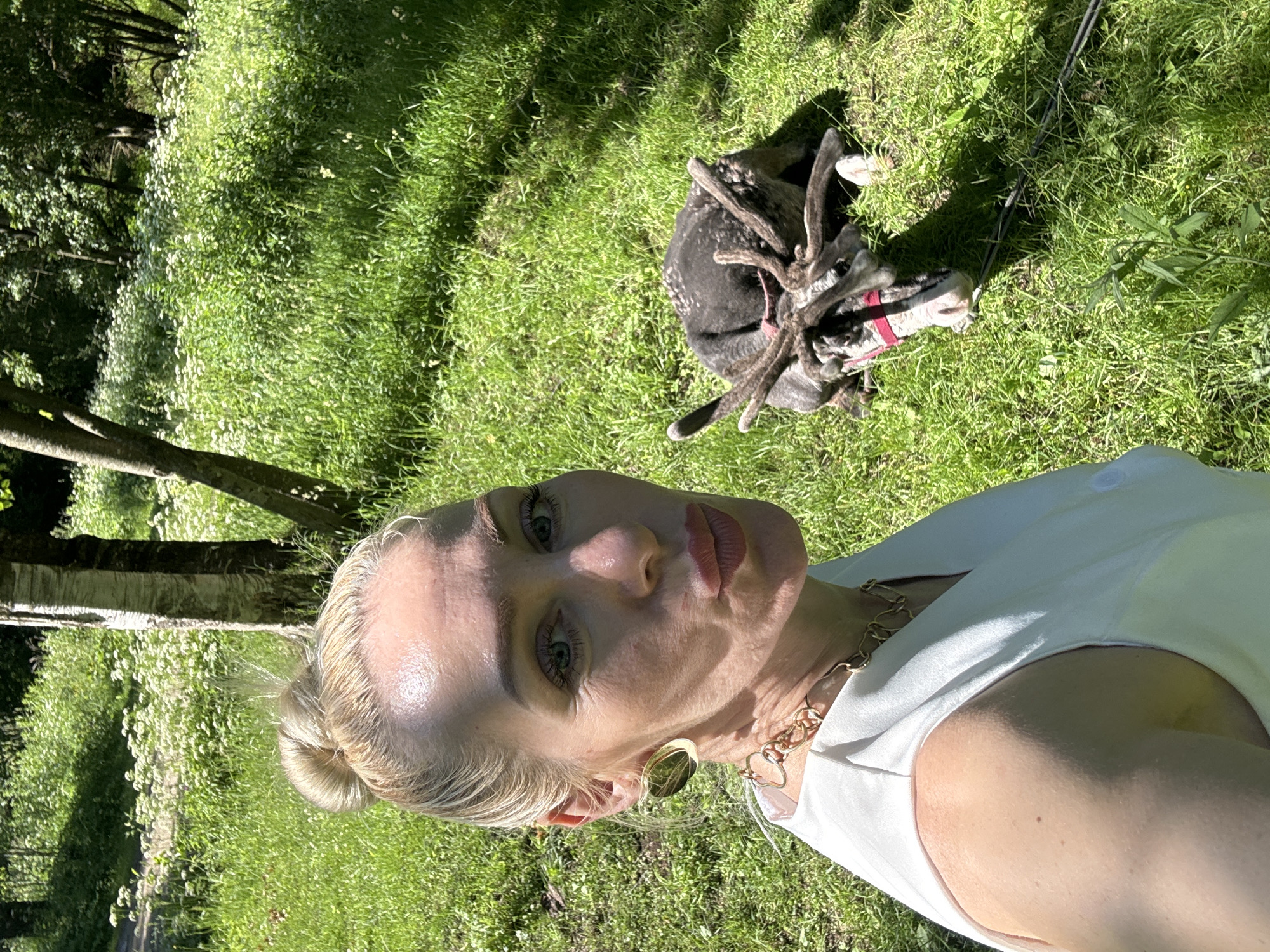 A woman with blonde hair in a bun and large gold hoop earrings, wearing a white top and a gold chain necklace, poses for a selfie with a reindeer lying on the grass behind her. The reindeer has antlers and a red halter. The scene is set outdoors in a grassy area with trees and white wildflowers in the background, likely within the city of Pavlovsk, Russia. The sunlight casts shadows on the ground, indicating a bright, sunny day. The woman is leaning in towards the camera, with her lips pursed. The reindeer appears to be resting peacefully.