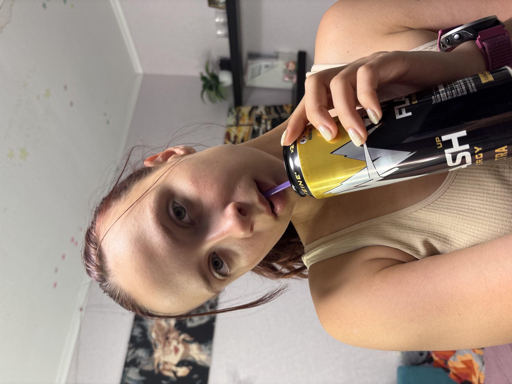 A young Caucasian female is depicted indoors, looking directly at the viewer while drinking from a tall black and yellow "FLASH UP ENERGY ULTRA" can via a purple straw. She has reddish-brown hair, light eyes, and wears a light beige ribbed tank top. A watch with a fuchsia fabric strap and dark face is on her right wrist. Her fingernails are painted a light, speckled color. Her right hand holds the energy drink can, which features a prominent white/grey lightning bolt design and visible text including "CAFFEINE" in both English and Cyrillic. The background reveals an indoor residential setting in the city of Прикамский, Russia. The white ceiling has faint, pastel star and dot decals in yellow, blue, and pink, with white crown molding meeting light purple walls. On the left wall, a large framed artwork depicts stylized female faces. On the right, two black floating shelves are mounted; the top shelf holds a small green potted plant and two clear glasses, while the bottom shelf has smaller, indistinct items. Behind the shelves, another piece of abstract art in warm tones is visible. Partial views of teal, orange, and muted-colored fabrics are visible in the lower left background.