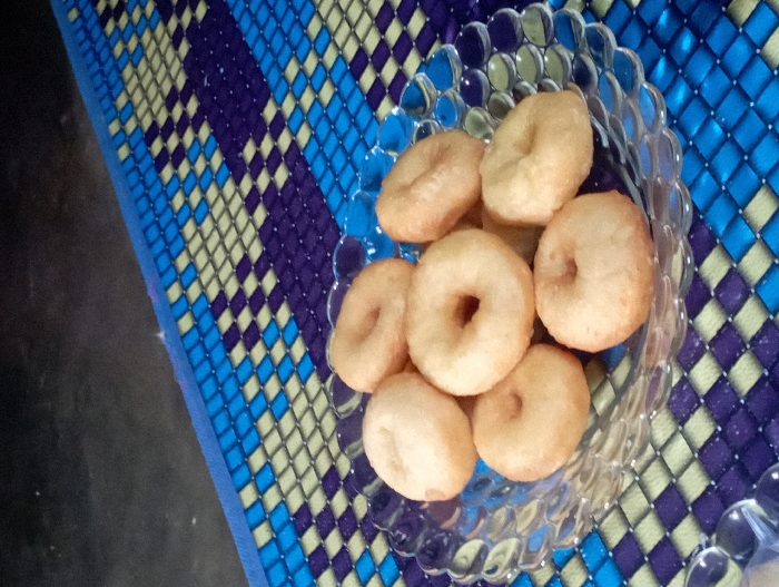 A clear, fluted glass bowl filled with approximately eight to ten small, golden-brown, ring-shaped pastries, resembling mini donuts, is the central focus of the image. The pastries have a slightly irregular, rustic appearance, suggesting they are freshly made.
The bowl rests on a colorful, patterned mat. The mat displays a geometric design of interlocking diamonds or squares in varying shades of blue (light and dark), deep purple, and a light yellow or cream color. The pattern runs diagonally across the frame, creating a dynamic background for the food. The texture of the mat suggests it is woven, possibly from plastic or a similar durable material.
To the left and underneath the mat, a dark, solid surface is visible, likely a floor or table, providing a somber contrast to the vibrant mat. The lighting in the image is soft and ambient, characteristic of an indoor setting, and does not suggest a specific time of day. No people are present, nor is any explicit activity taking place; the scene simply presents the prepared food. There is no visible text in the image. The setting suggests a casual, domestic environment, possibly in Pubian, Indonesia, given the context.