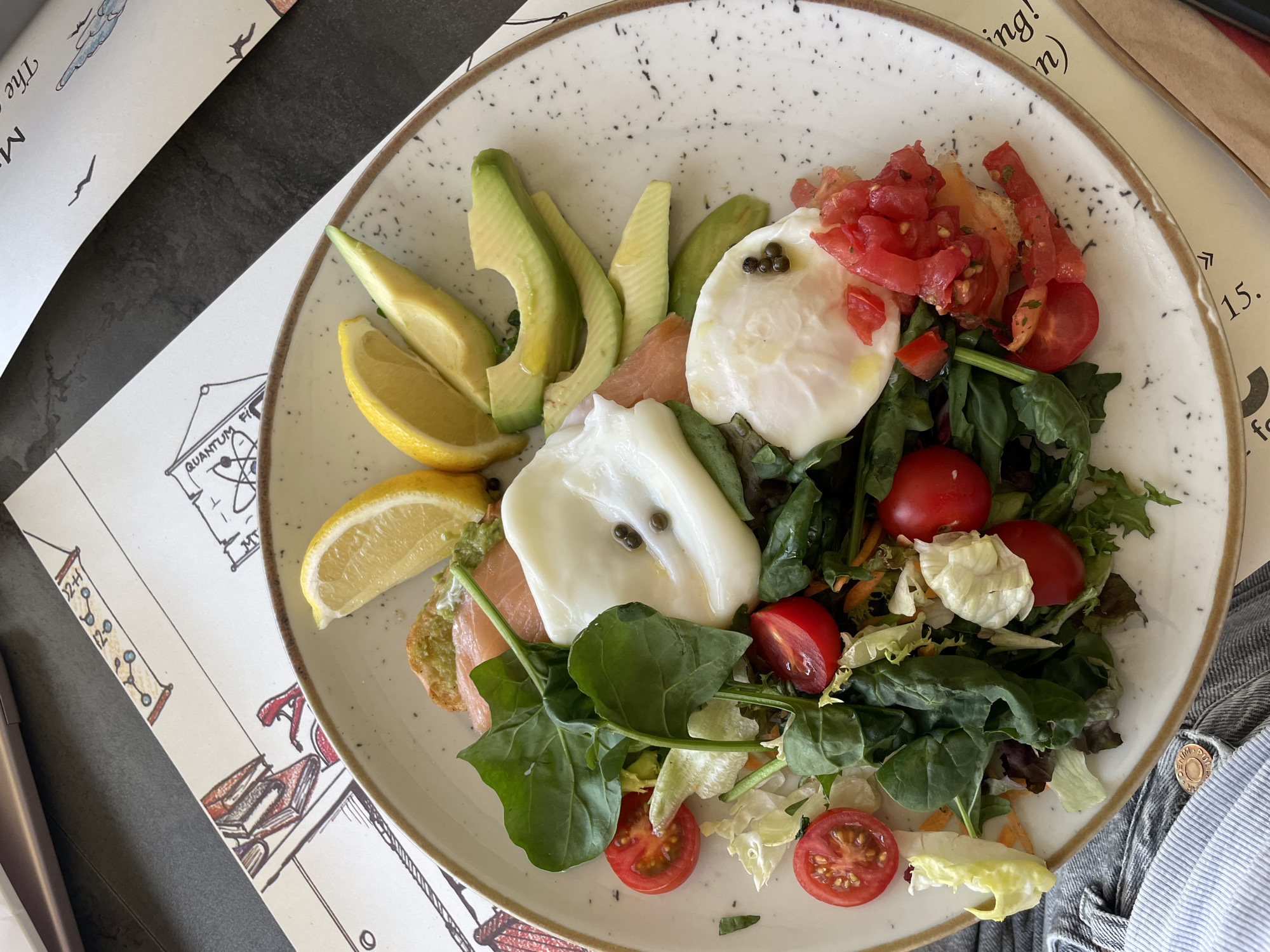 A close-up, overhead shot captures a plate of food and a portion of a table setting. The plate contains a vibrant salad composed of mixed greens, halved cherry tomatoes, sliced avocado, smoked salmon, and two poached eggs, each adorned with capers. Lemon wedges are placed on the side. The plate rests on a table with a patterned placemat featuring line drawings of books and scientific diagrams, along with some text. A folded piece of paper with text and a paper coaster are also visible on the table. The background is dark and slightly out of focus. The lighting is bright, suggesting daylight. No specific location details like Budva, Montenegro are present in the visual information provided.